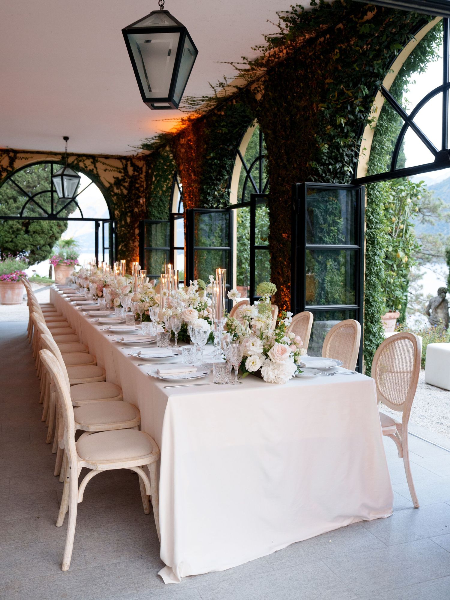 Long banquet table with white linens and floral arrangements set against arched windows and climbing vines in an elegant venue.