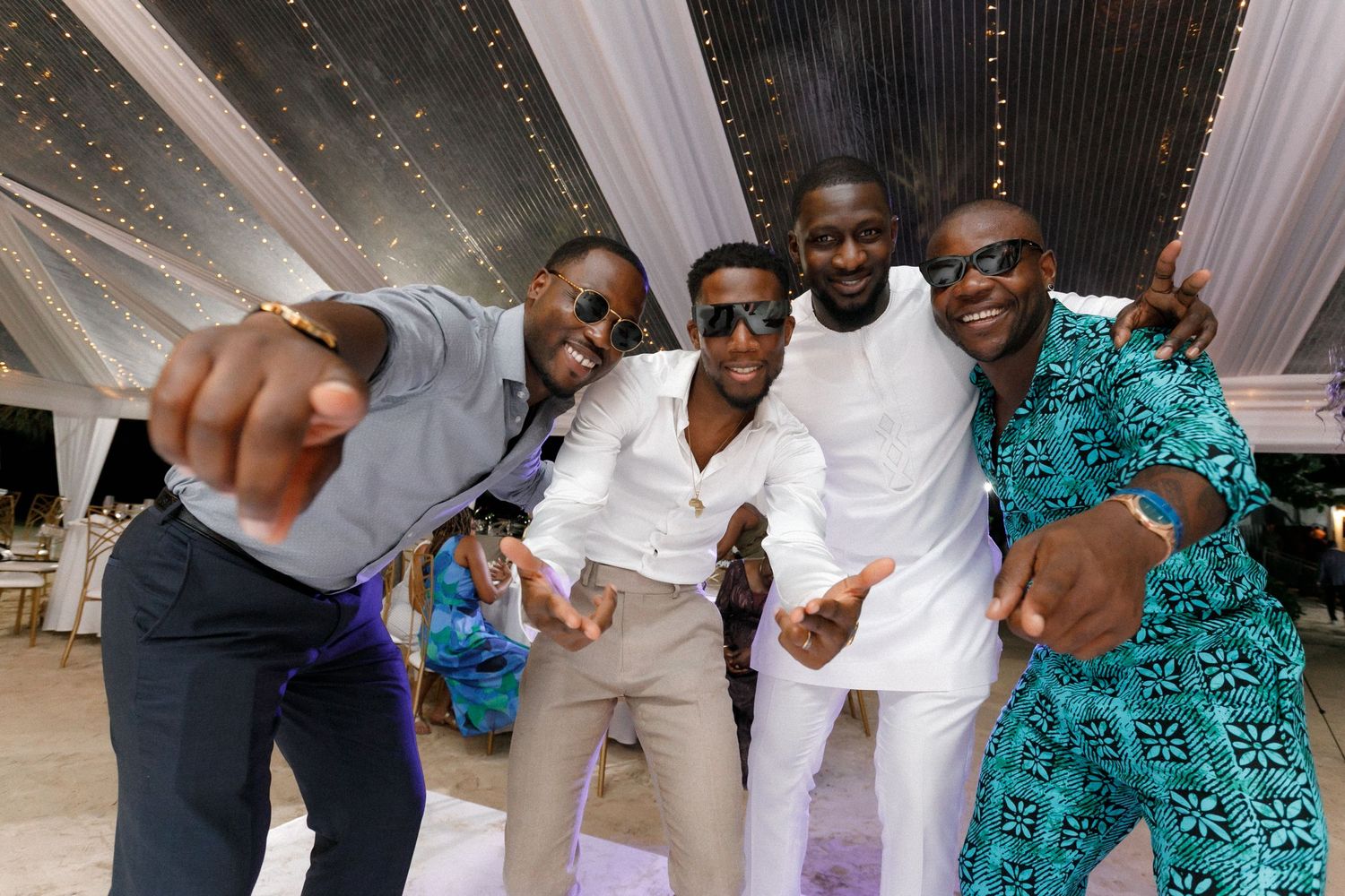 Wedding guests in stylish outfits pose playfully for a group photo under decorative lighting.