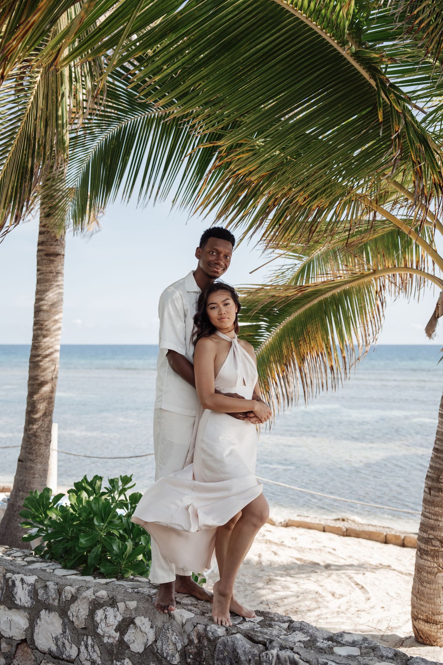 Couple in white outfits posing between palm trees at tropical beach location.