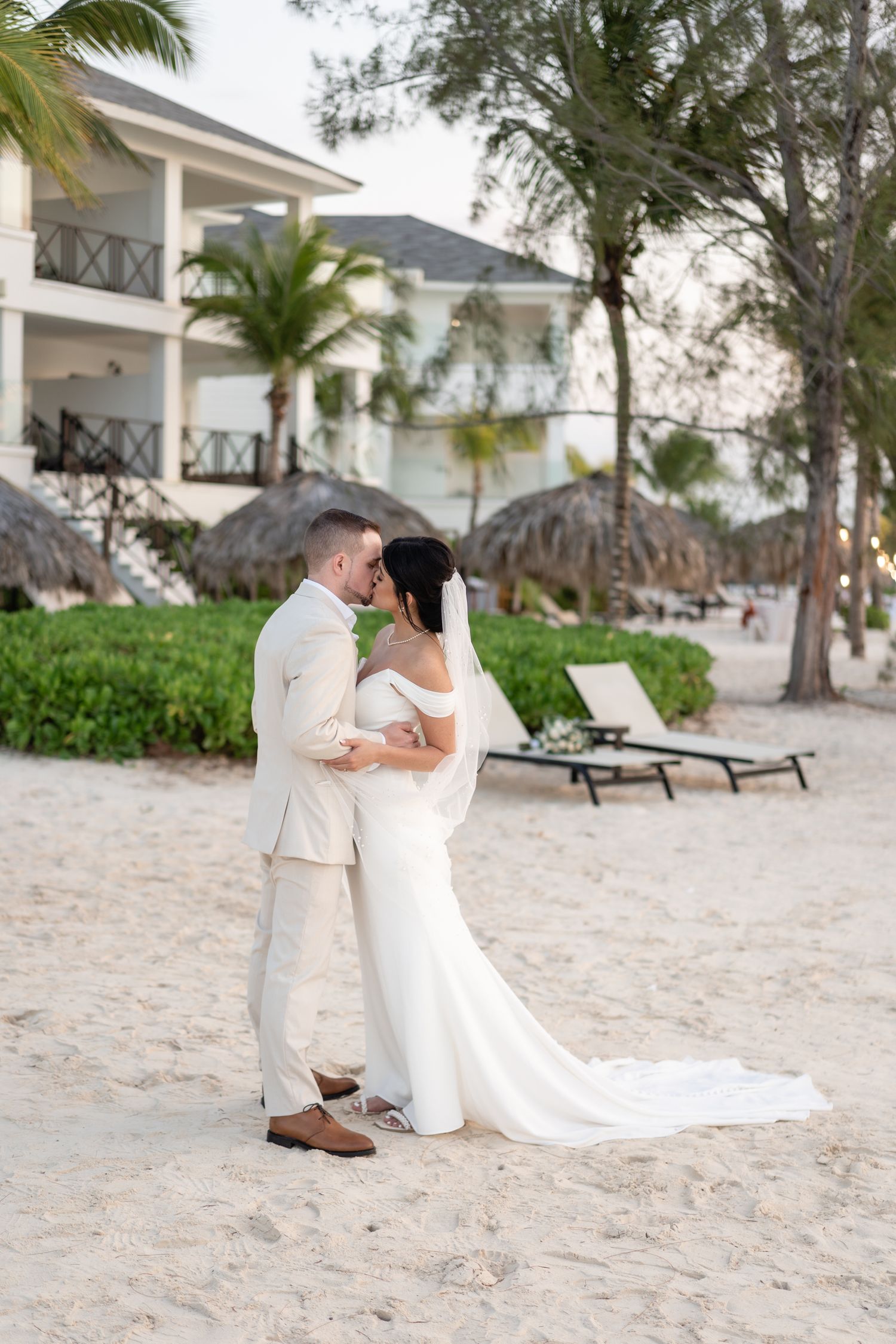 Destination beach wedding couple embracing near palm trees and resort buildings.