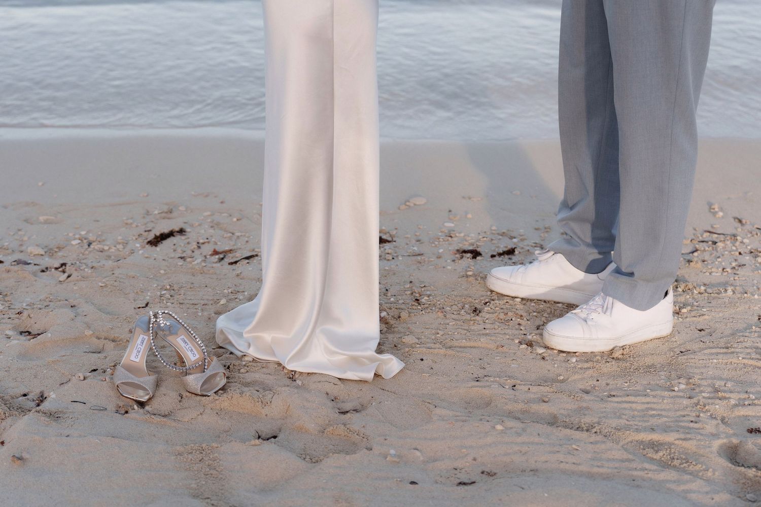 Close-up of formal shoes and dress hems on sandy beach.