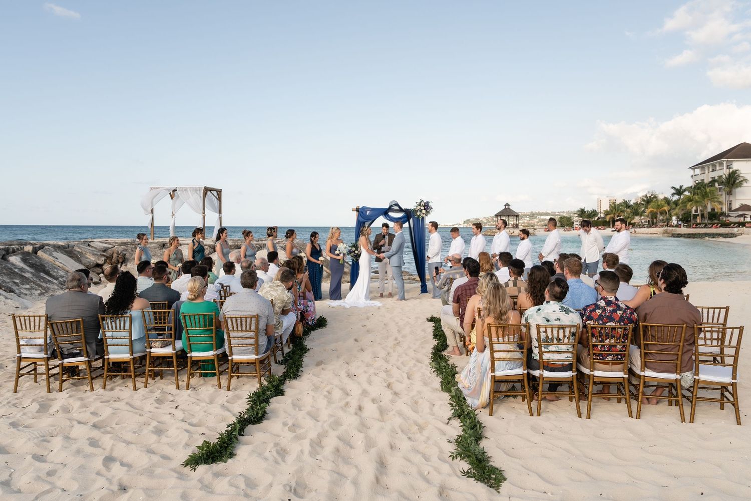Beach wedding ceremony setup with wooden chairs and greenery-lined aisle facing the ocean.