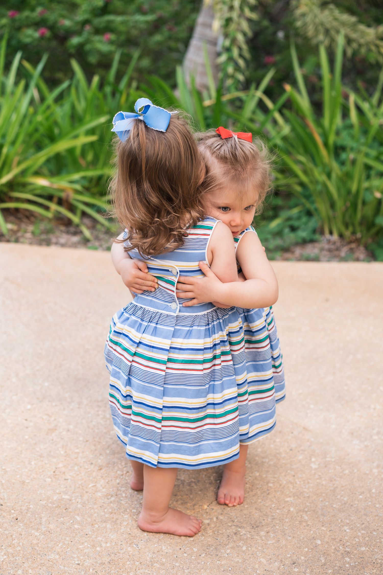 Two young children in matching striped dresses share a sweet hug on a garden pathway.
