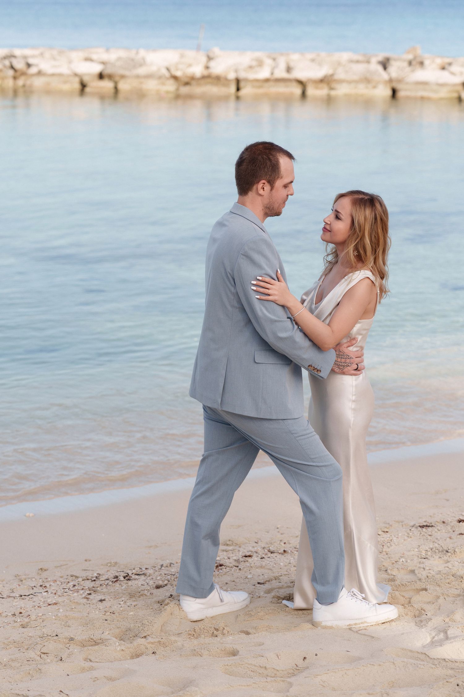 Couple in formal attire dancing together on a beach with rocky coastline in background.