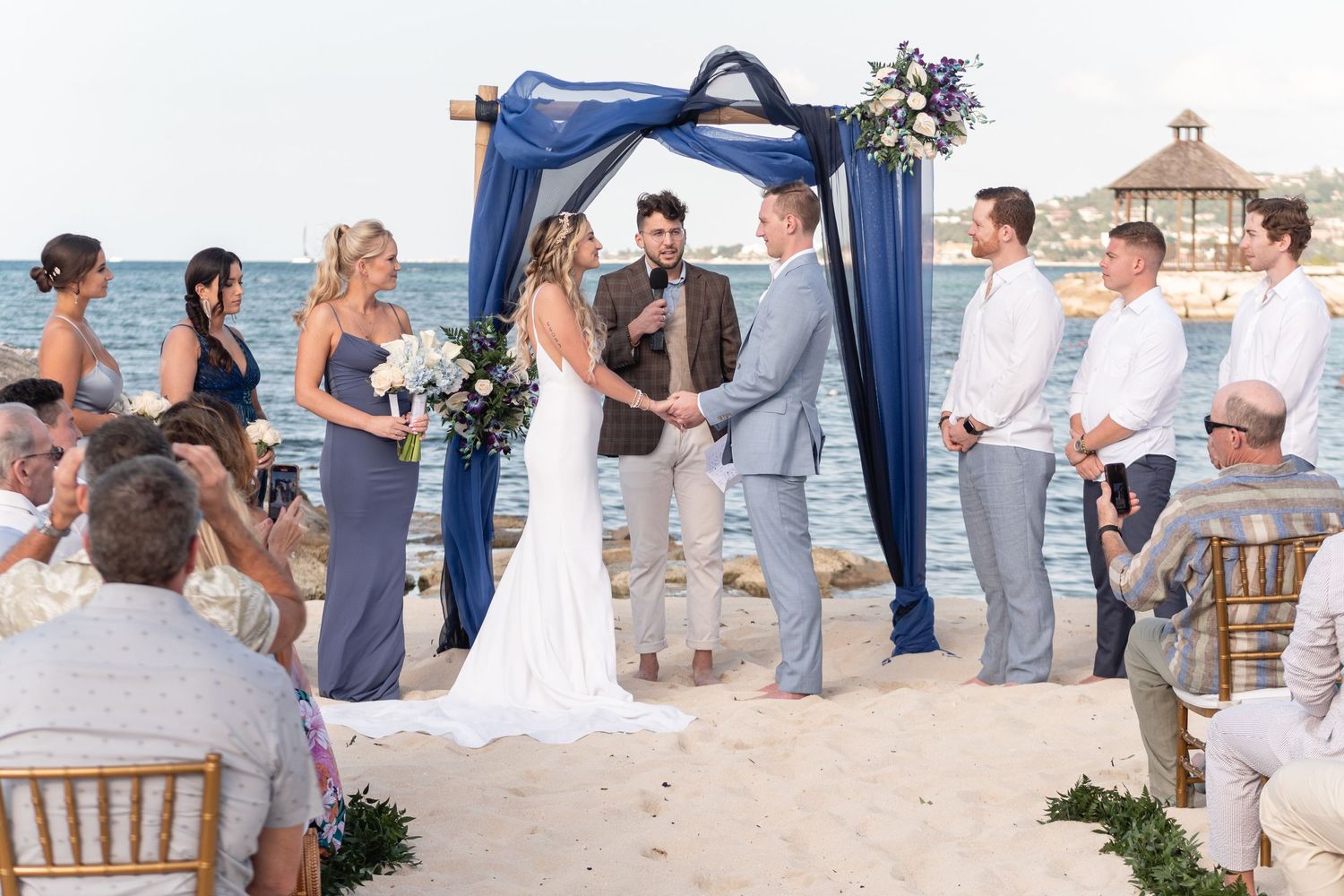 Beach wedding ceremony under blue fabric arch with guests seated on wooden chairs.