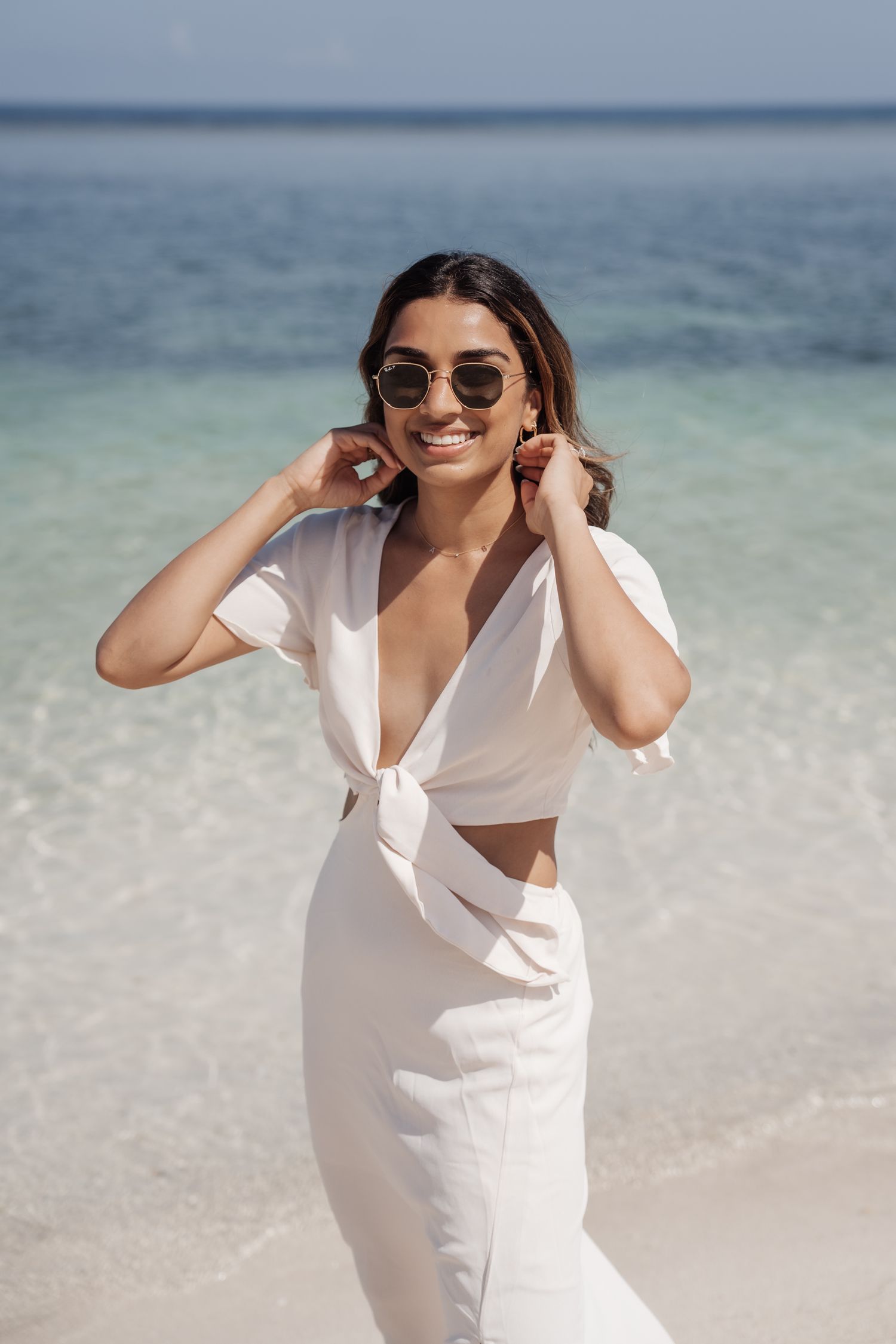 Woman in white cutout dress poses on beach wearing sunglasses.