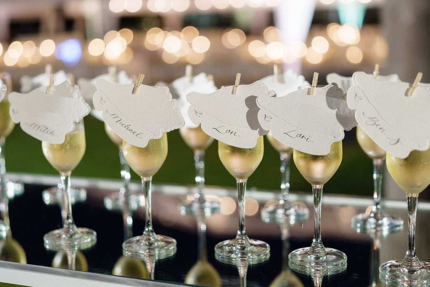Row of elegant cocktail glasses with white floral garnishes lined up on a mirrored surface with bokeh lights in background.