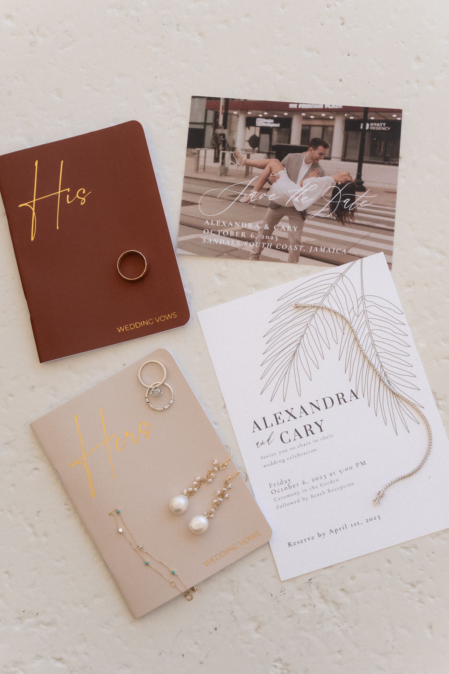 Wedding stationery flat lay with invitation, vow books, rings and jewelry on beige surface.
