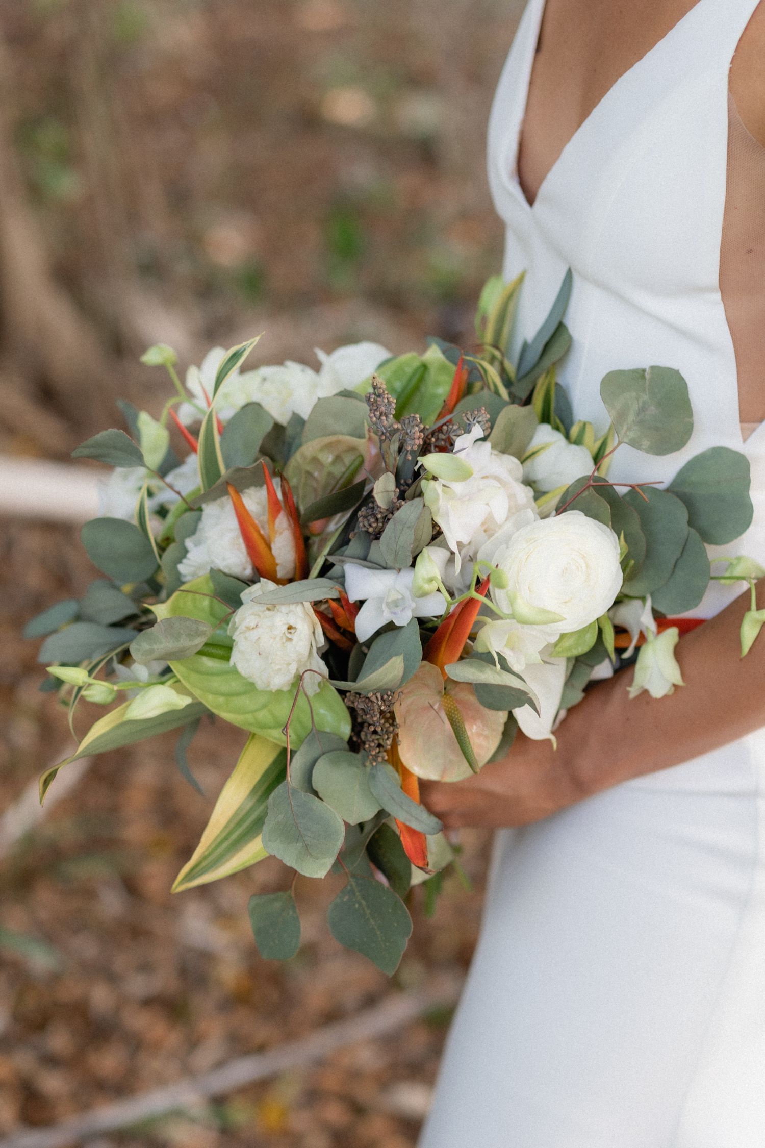 A wedding bouquet featuring white roses, eucalyptus, and orange tropical flowers against a white dress.