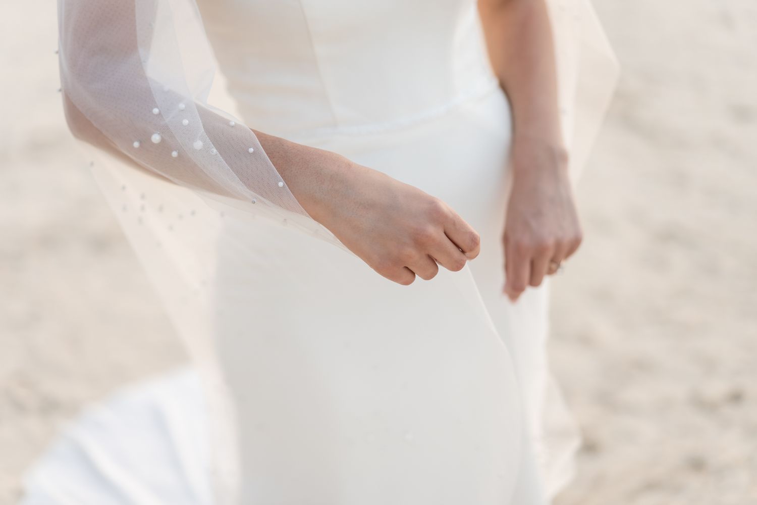 Close-up detail of a flowing white wedding dress against sandy beach background.