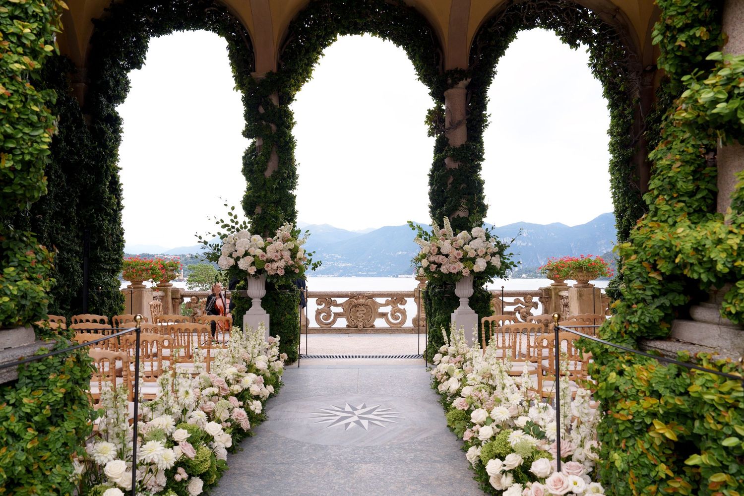 Scenic garden terrace with cypress trees and white roses lining a stone balustrade overlooking mountain views.