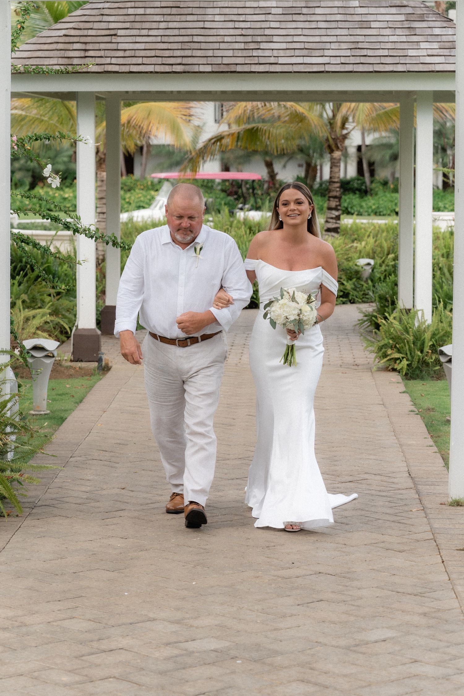 A bride in white dress walks down a covered pathway with her escort in light summer attire.