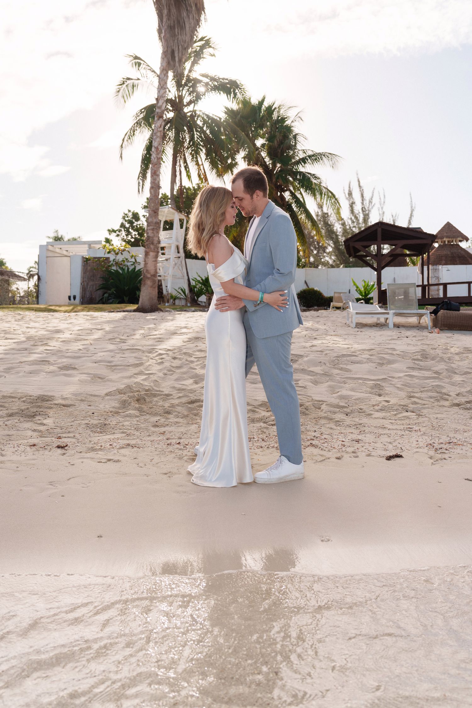 Couple in formal attire sharing intimate moment on tropical beach with palm trees.