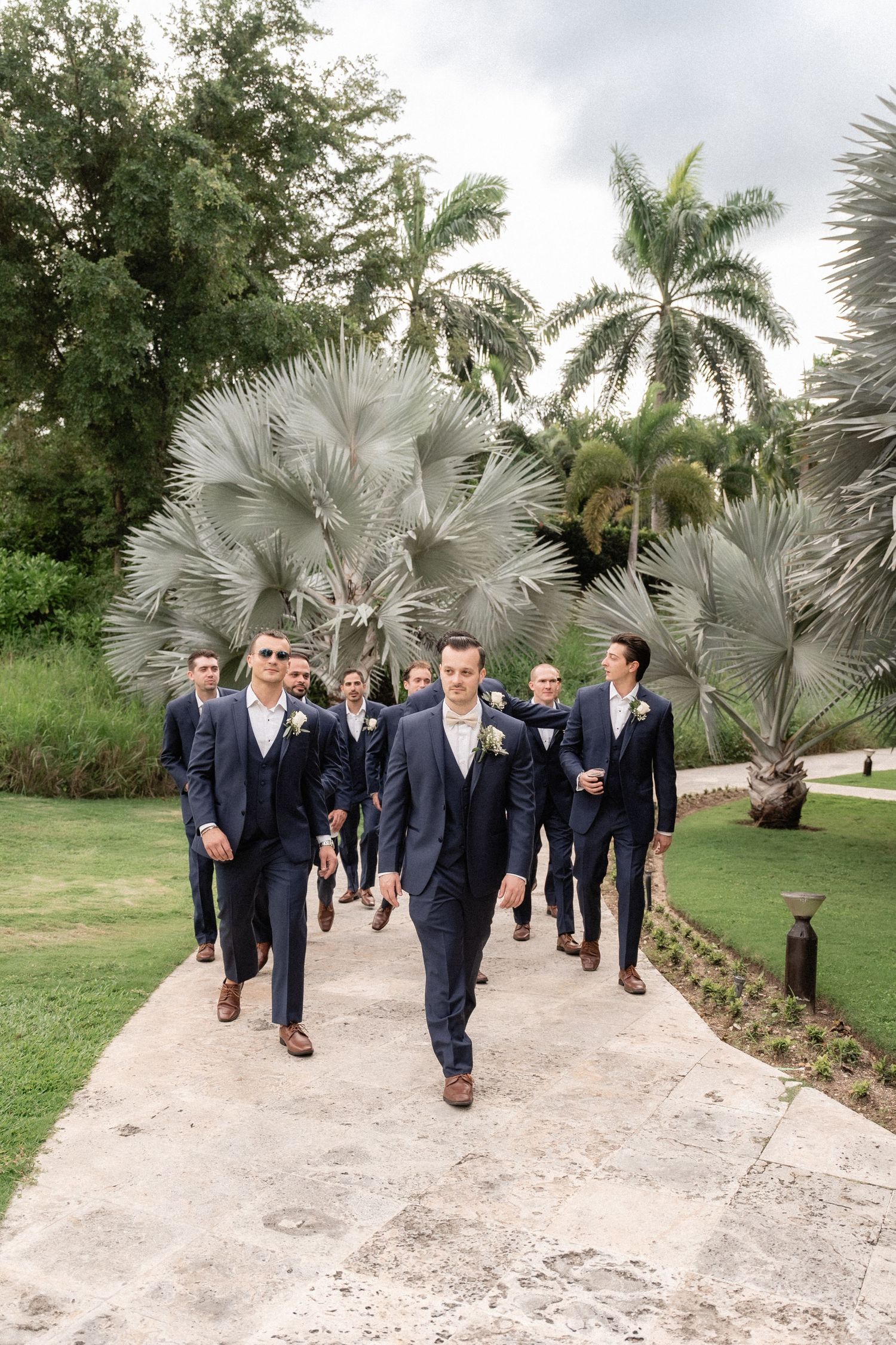 A group of groomsmen in navy suits walk along a palm-lined pathway.