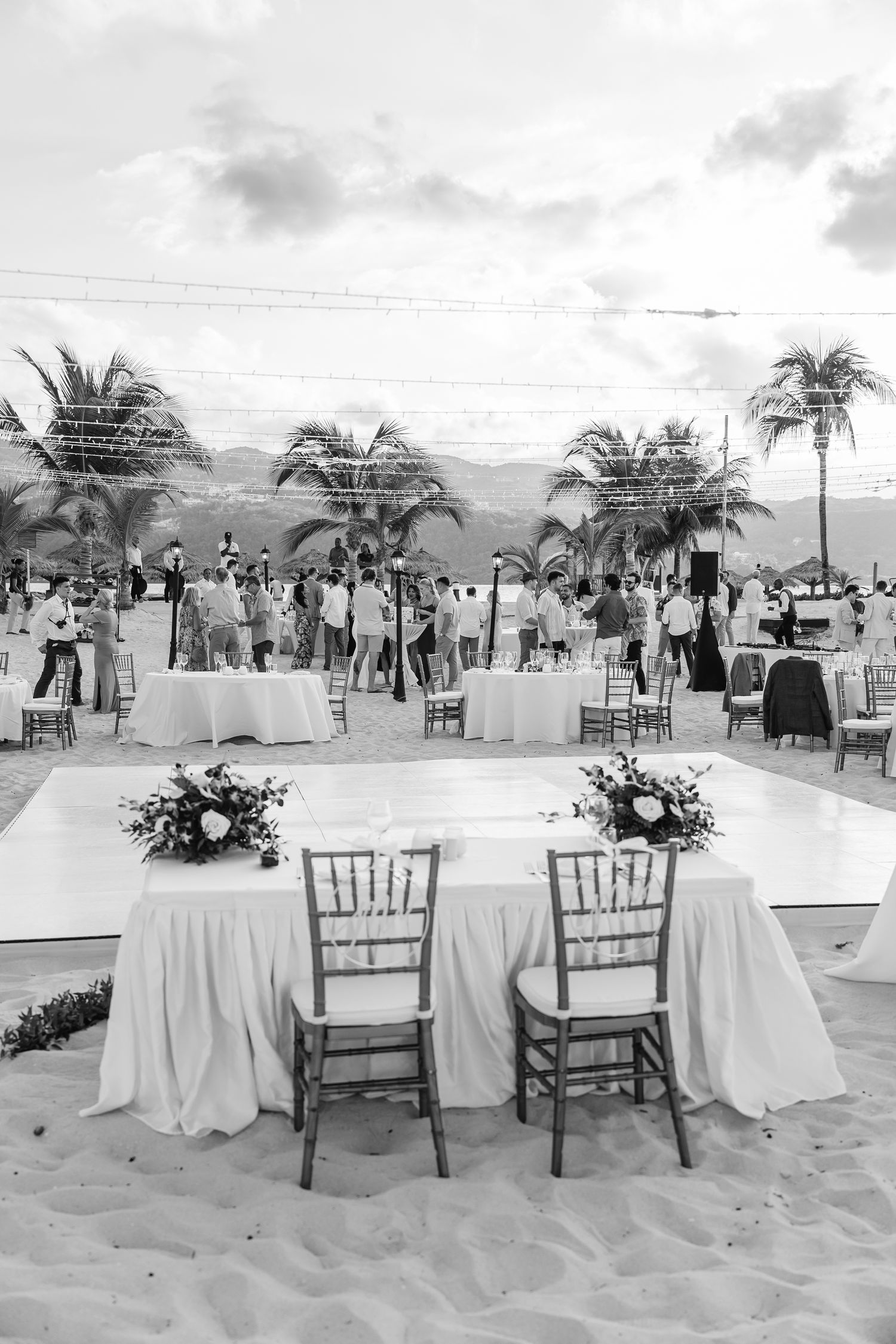 Black and white photo of a beach wedding reception setup with tables and palm trees.
