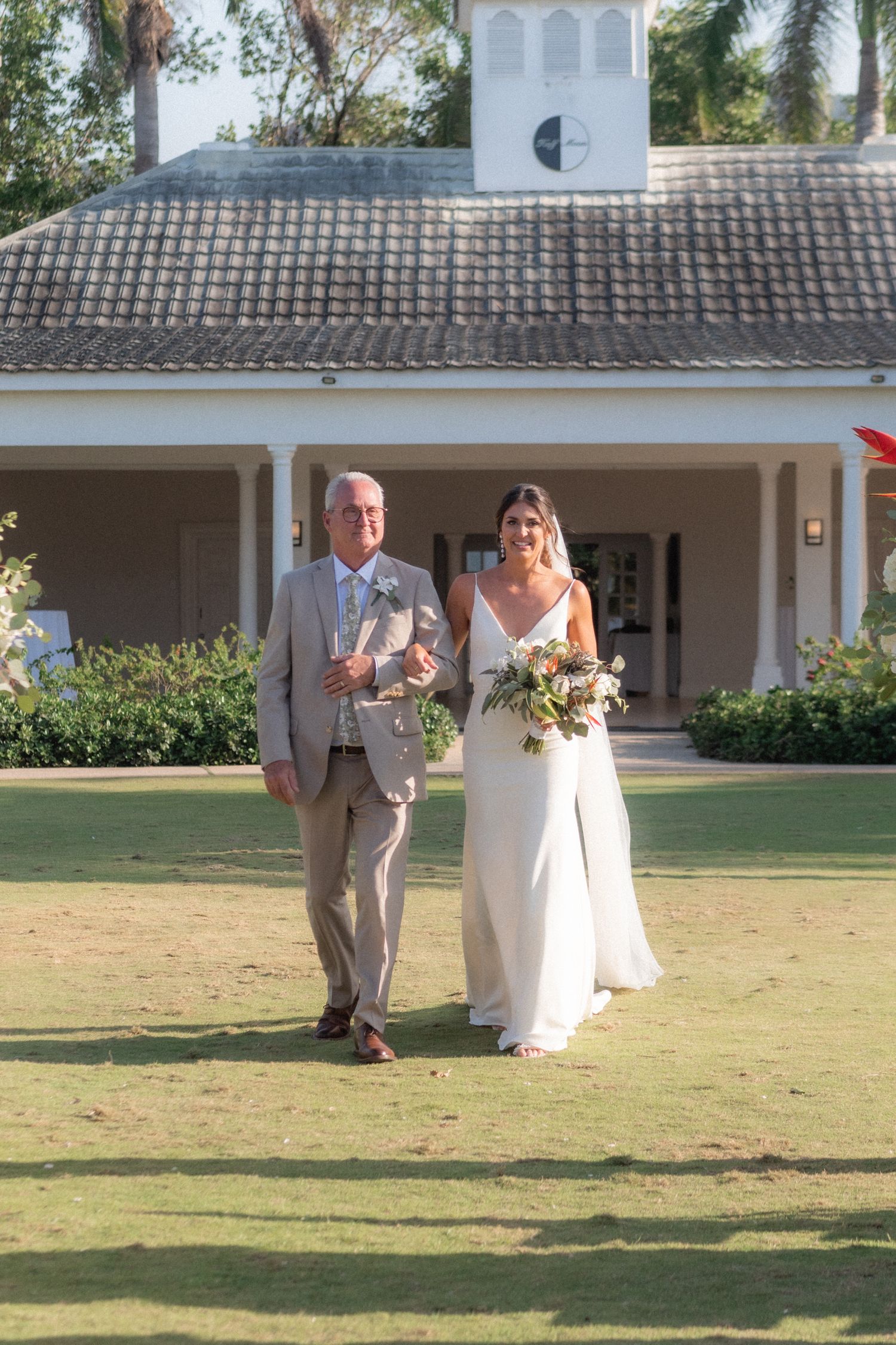 A wedding ceremony entrance at an elegant white colonial-style venue with a tiled roof and manicured lawn.