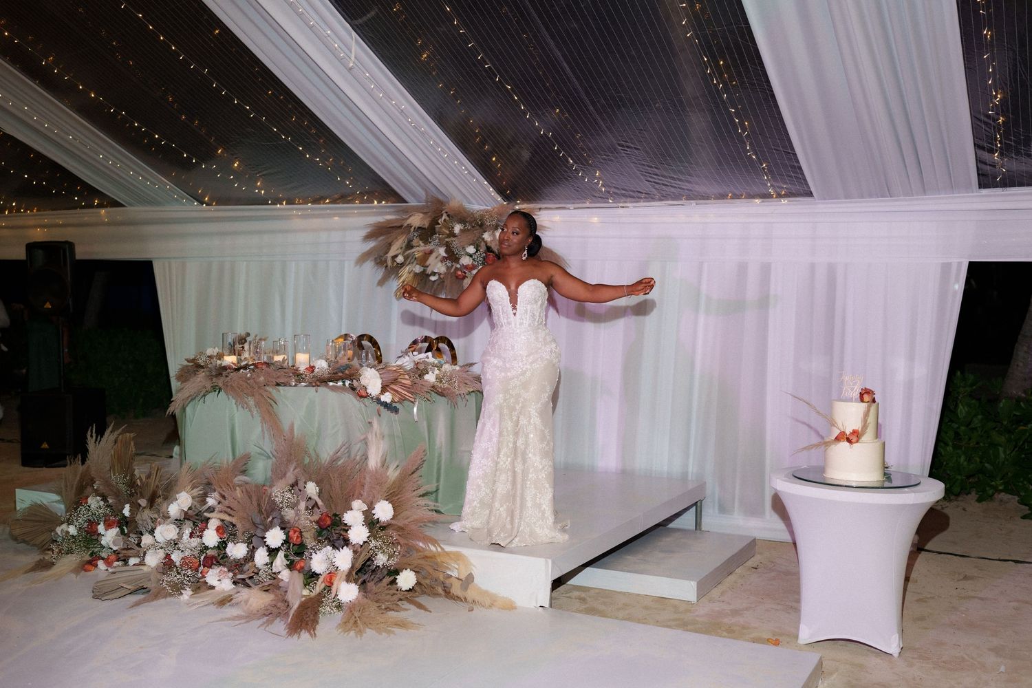 A bride poses on an elevated platform surrounded by elegant floral arrangements and white draping.