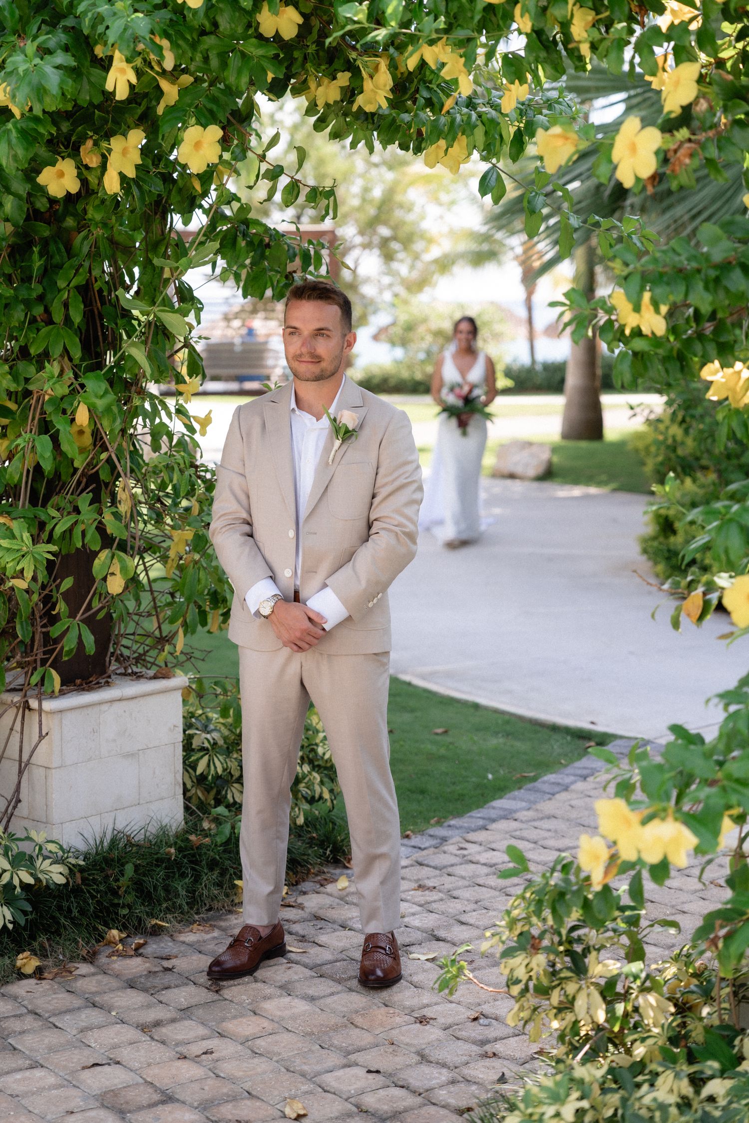 Groom in light beige suit stands on garden path with yellow flowering trees.