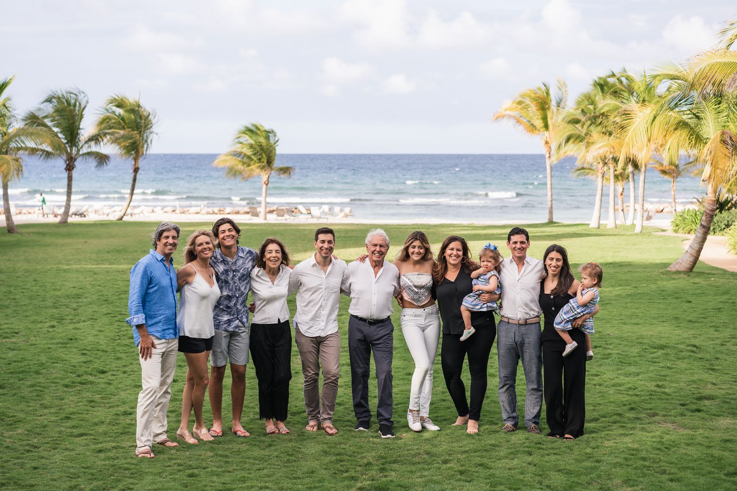 Wedding party poses for a group photo on a tropical lawn with palm trees and ocean view.