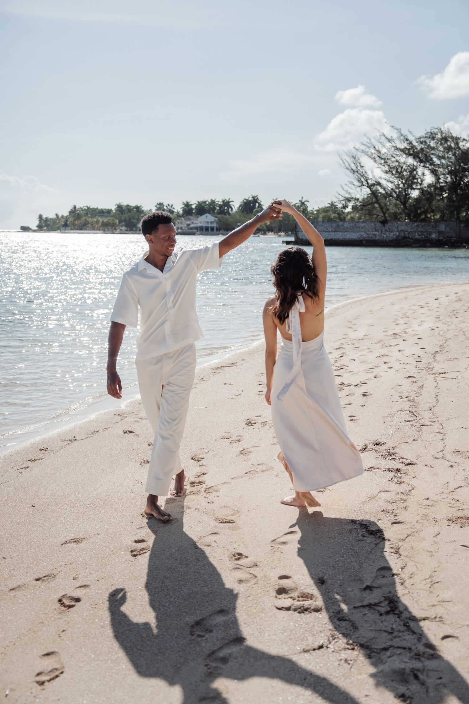 Couple in white dancing on beach with dramatic shadows cast on sand.
