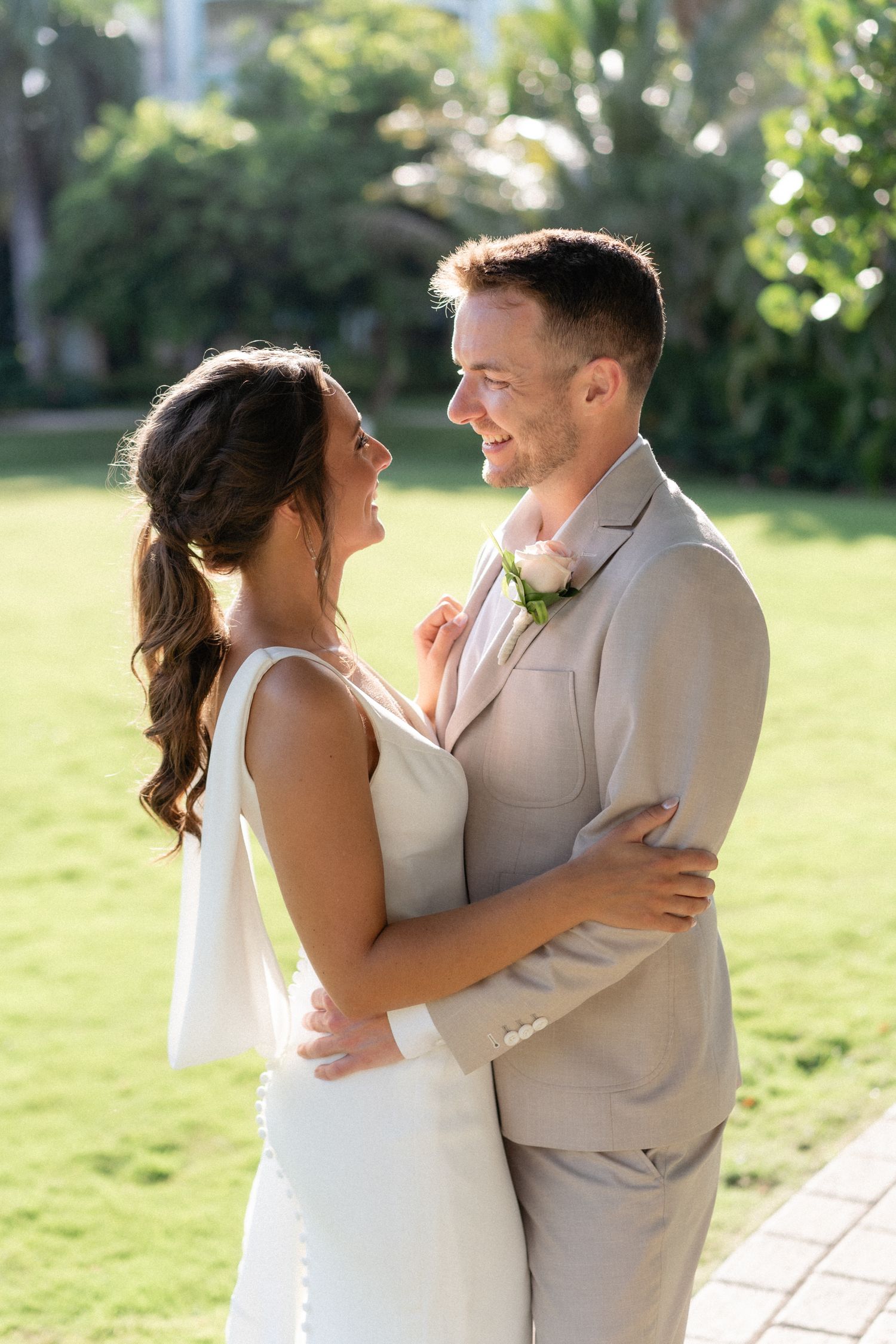 A newlywed couple embraces outdoors in formal attire against a garden background.