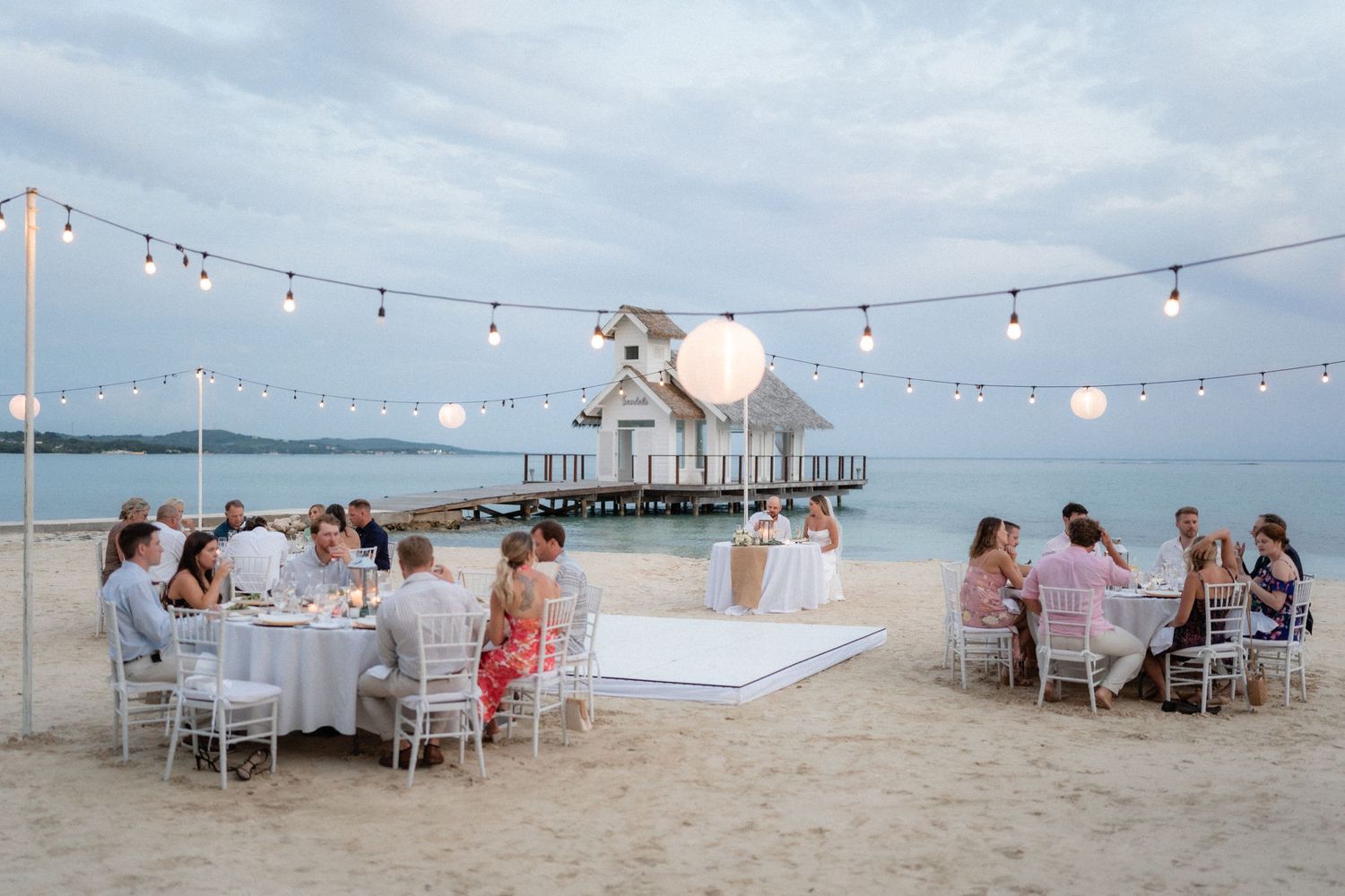 Beach wedding reception with string lights and guests dining at tables near ocean view.