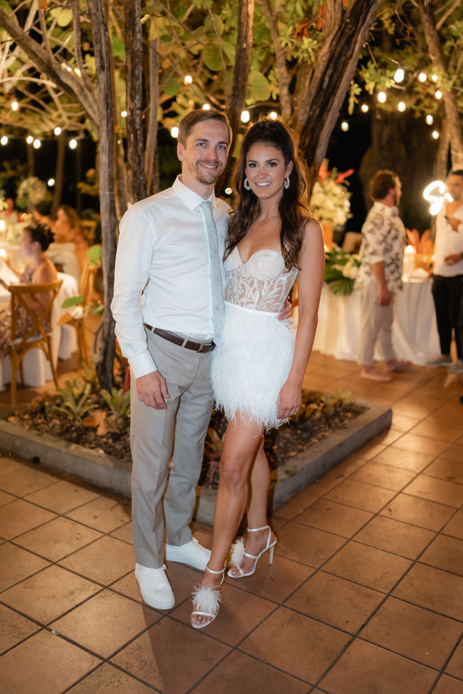 Newlyweds pose together at outdoor reception with string lights and tropical landscaping.