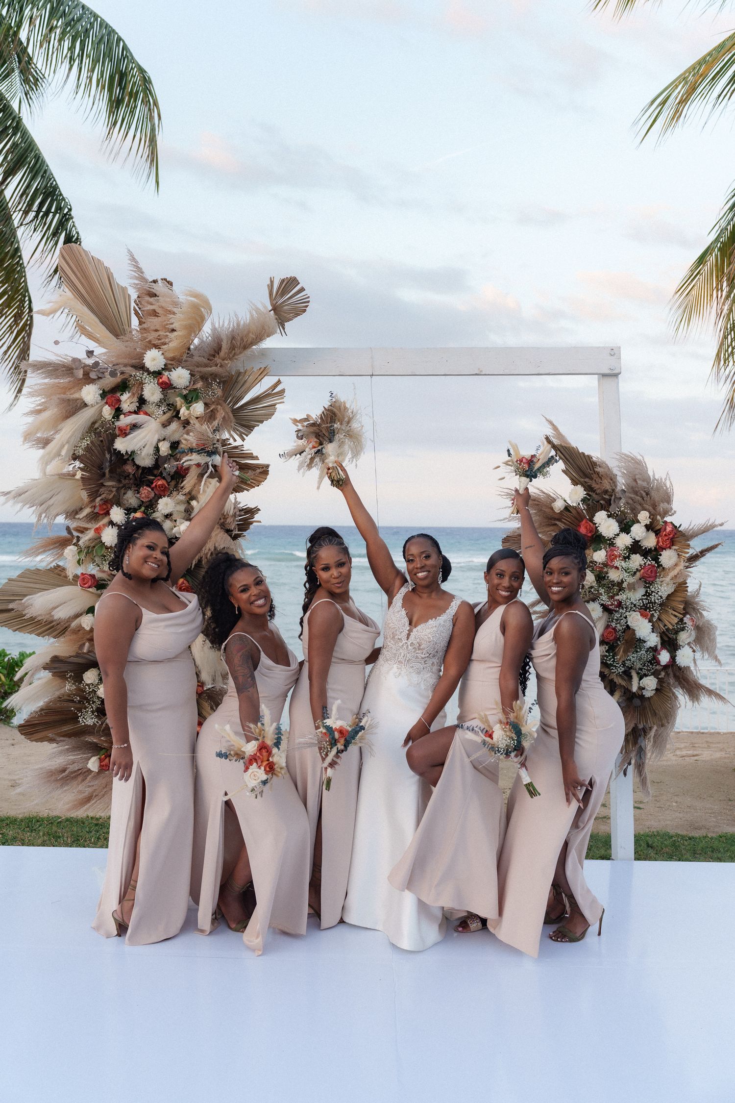 Bridesmaids in blush dresses pose by floral arch on oceanfront white platform.