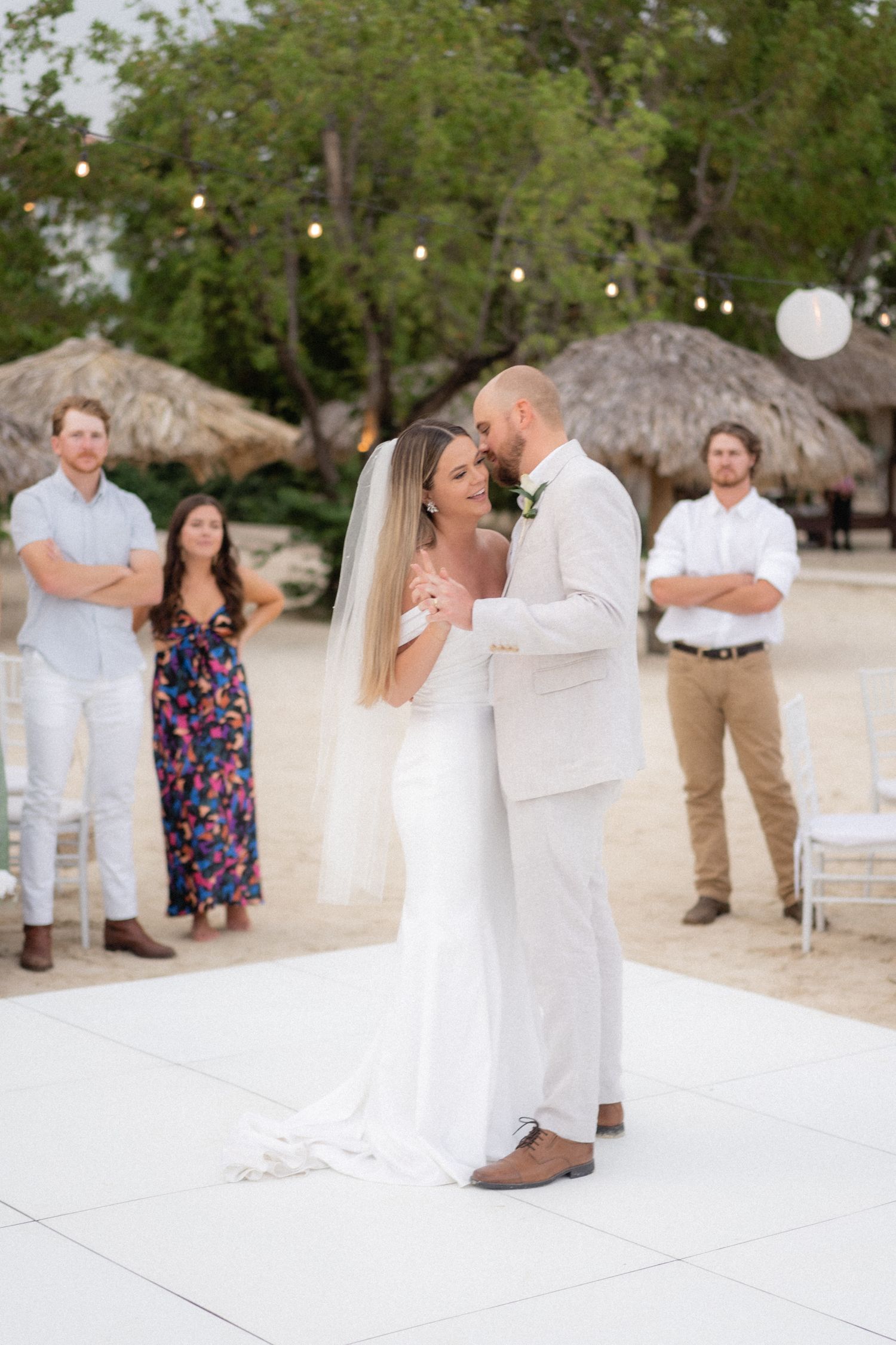 First dance at beach wedding reception with tiki huts and string lights in background.