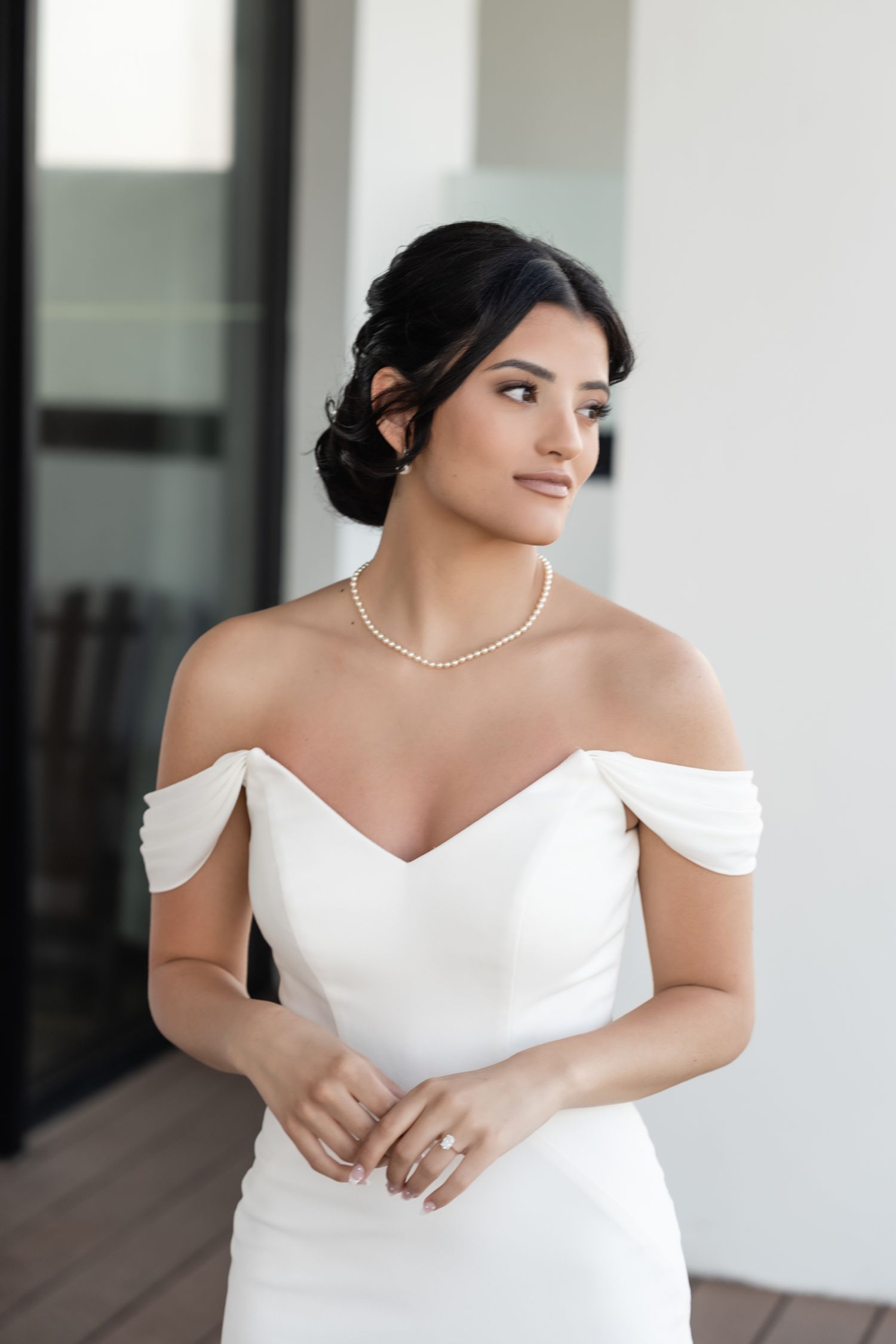 An elegant bride in an off-shoulder white dress and pearl necklace poses gracefully.