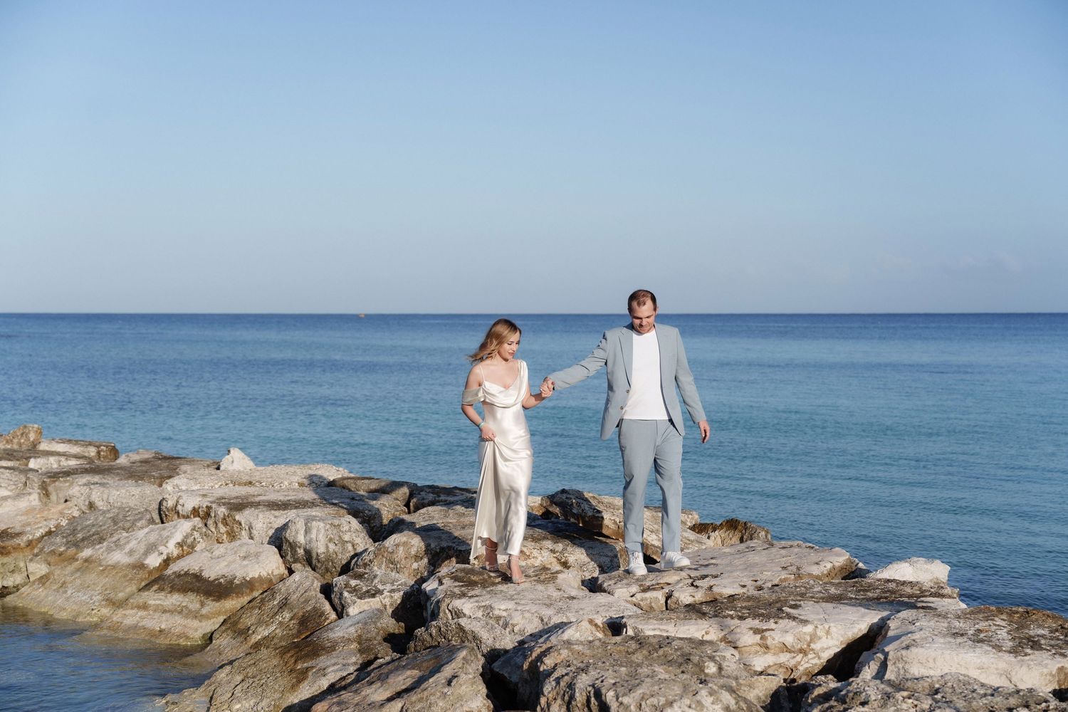 A couple in white and grey outfits standing together on rocky coastal formation.