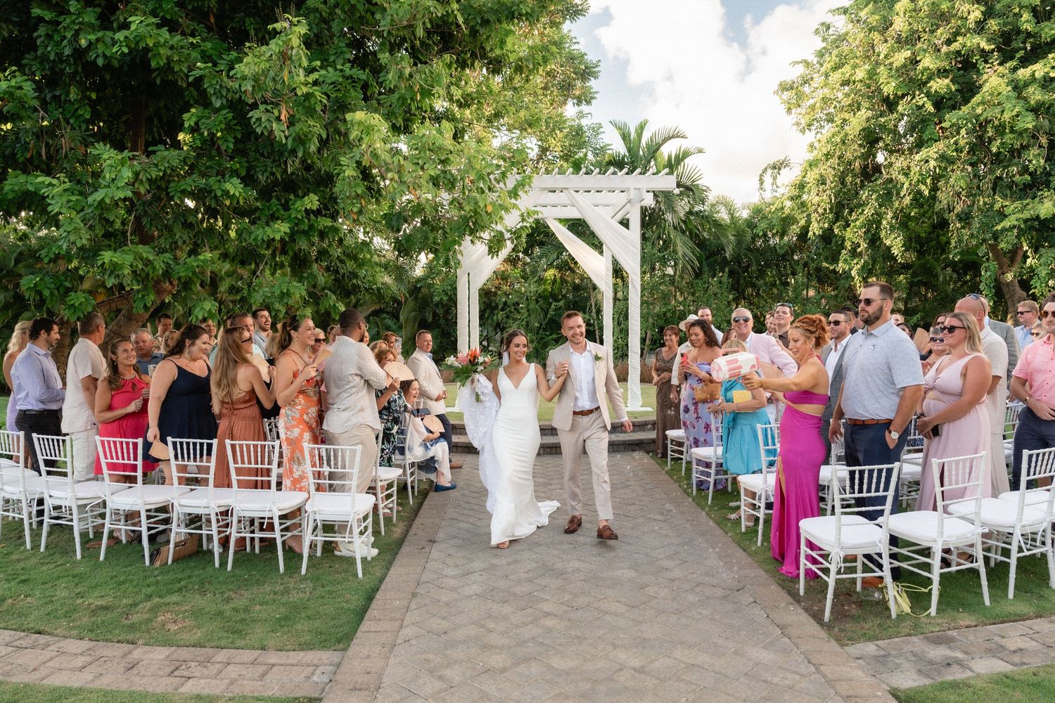 Newlyweds walk down the aisle while guests applaud at an outdoor garden ceremony.