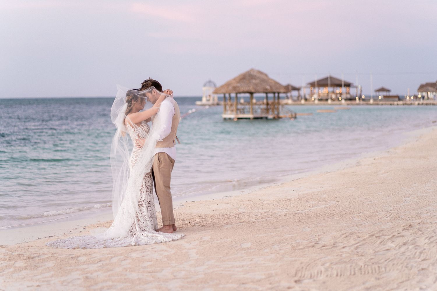 A romantic beach wedding photo with overwater bungalows visible in the distance at sunset.