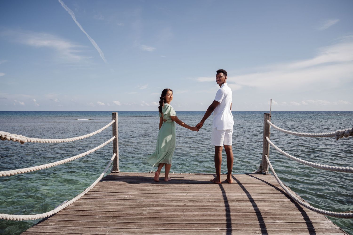 A couple holds hands while standing on a wooden pier over turquoise ocean waters.
