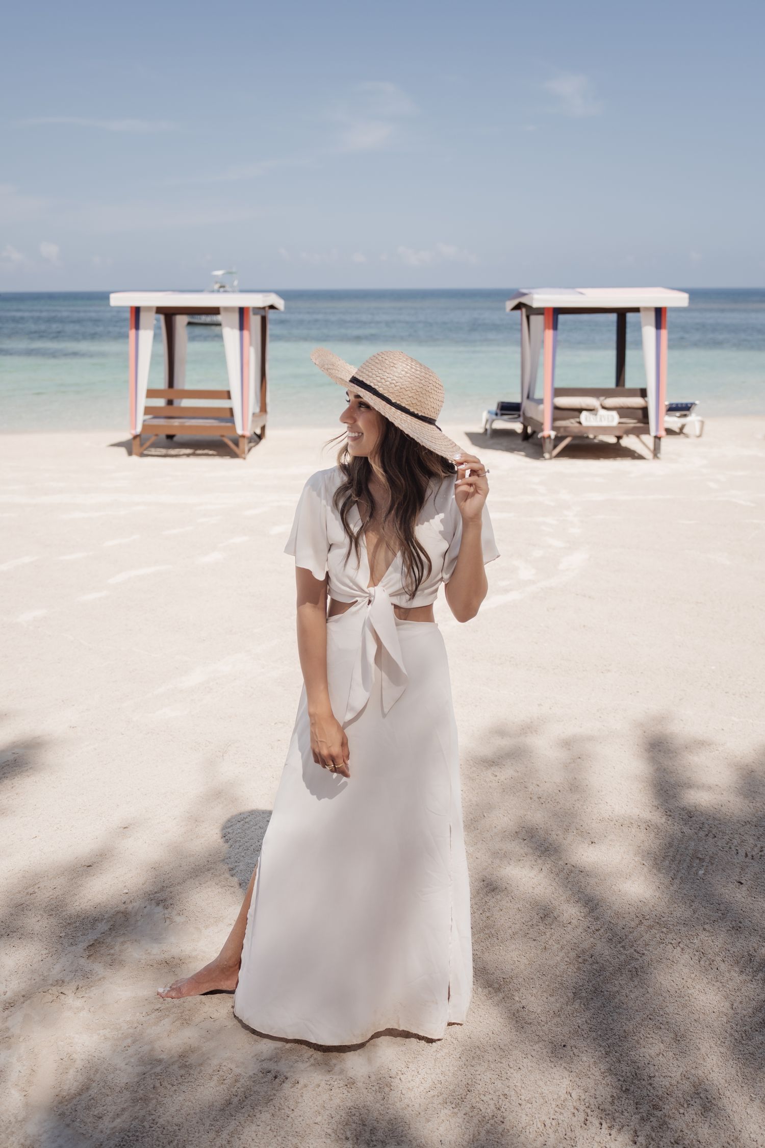 Woman in white dress and sun hat standing on beach with rustic cabanas in background.