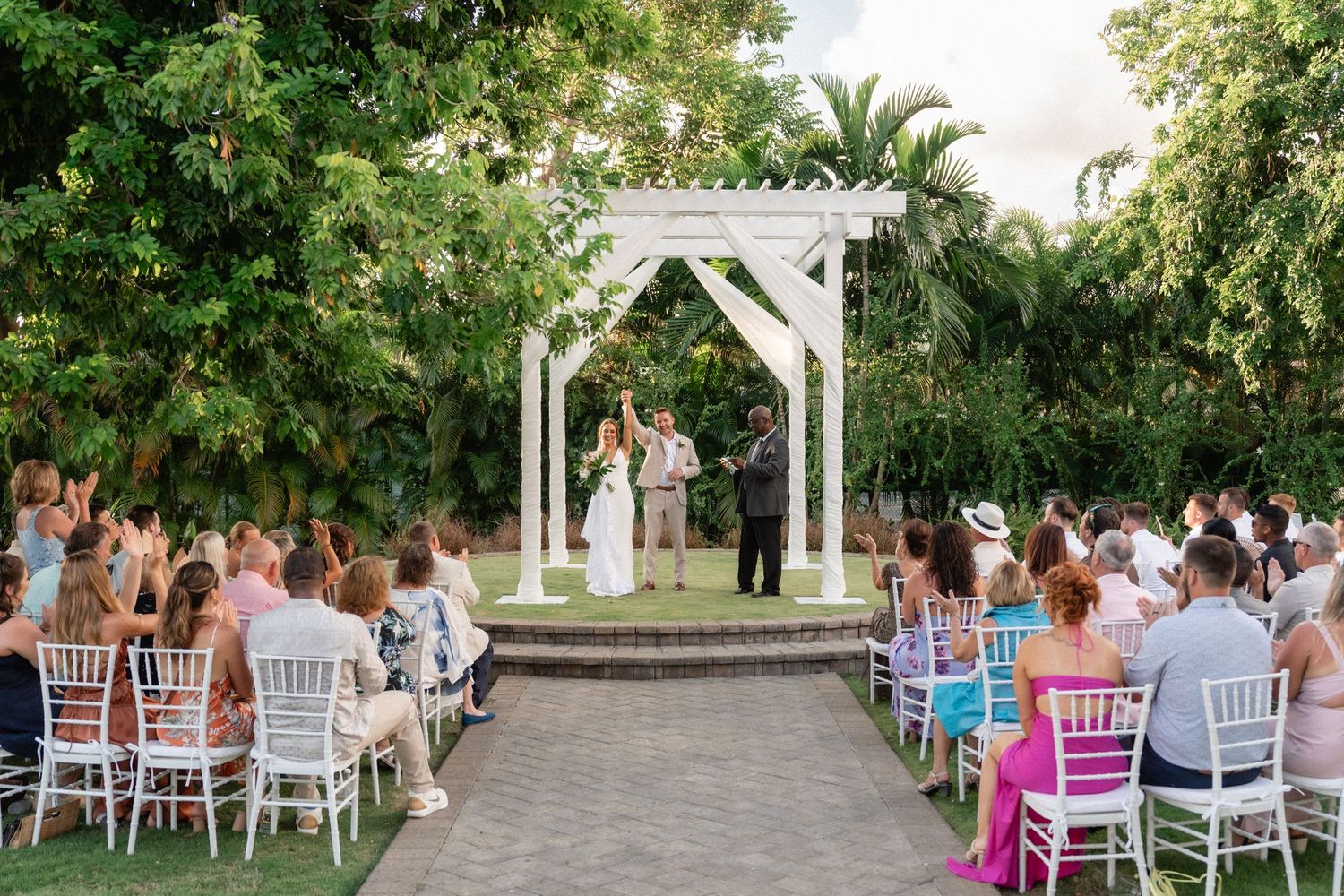 Wedding ceremony setup with white pergola altar and rows of chairs on stone pathway surrounded by greenery.