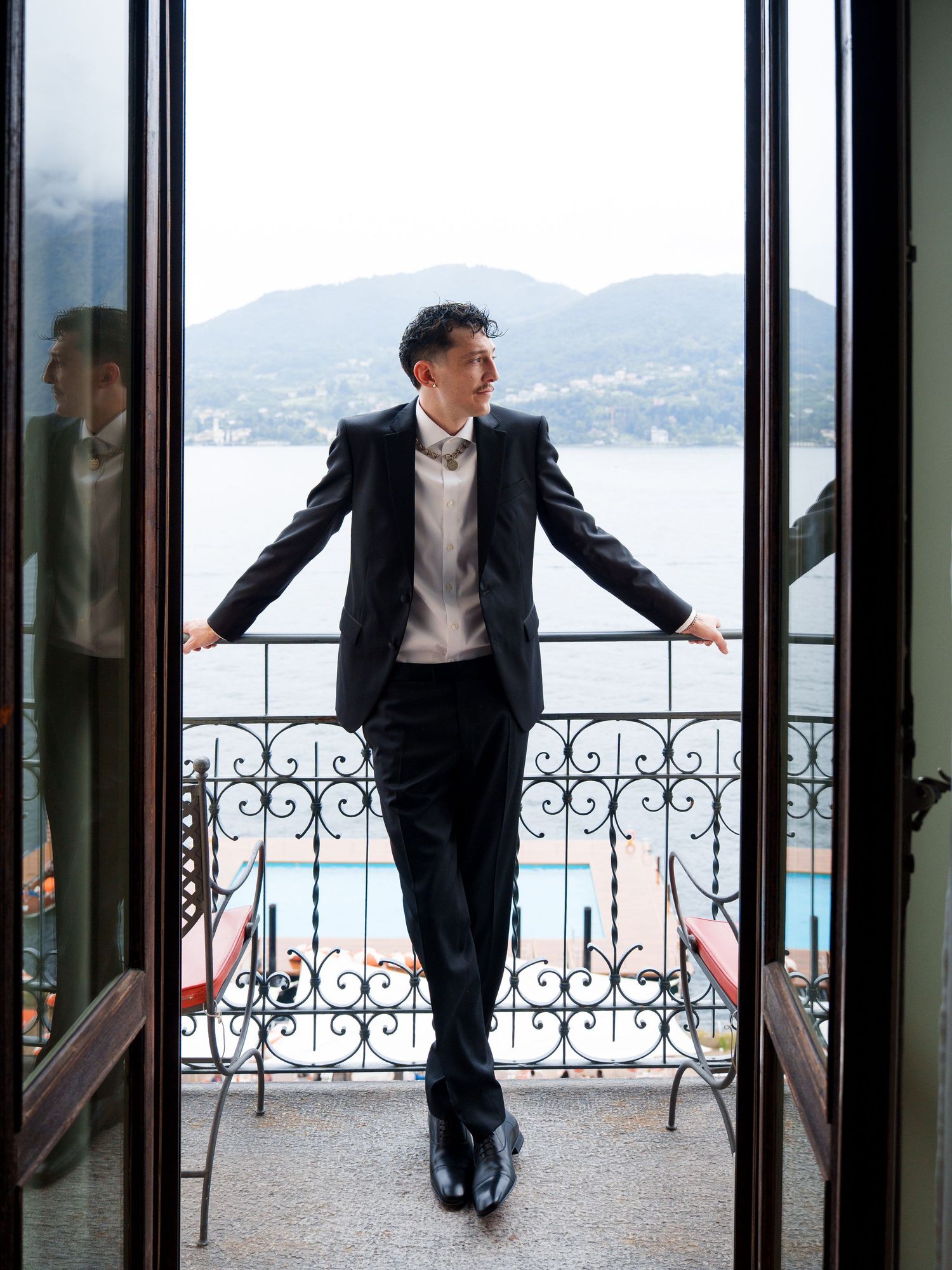 A person in a dark suit stands on an elegant balcony overlooking a scenic mountain lake view through ornate doors.