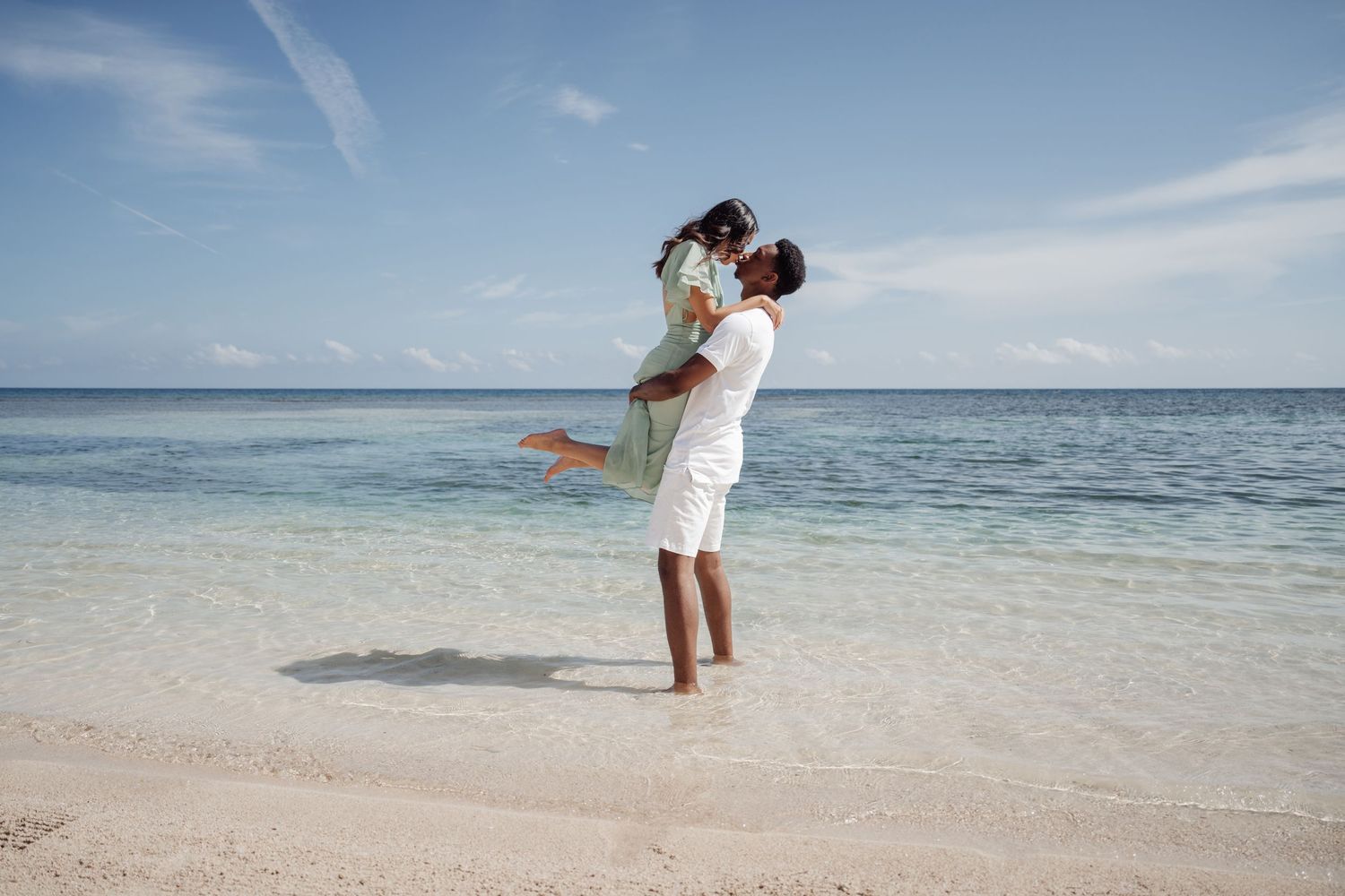 Playful beach photo with one person lifting the other against bright ocean backdrop.