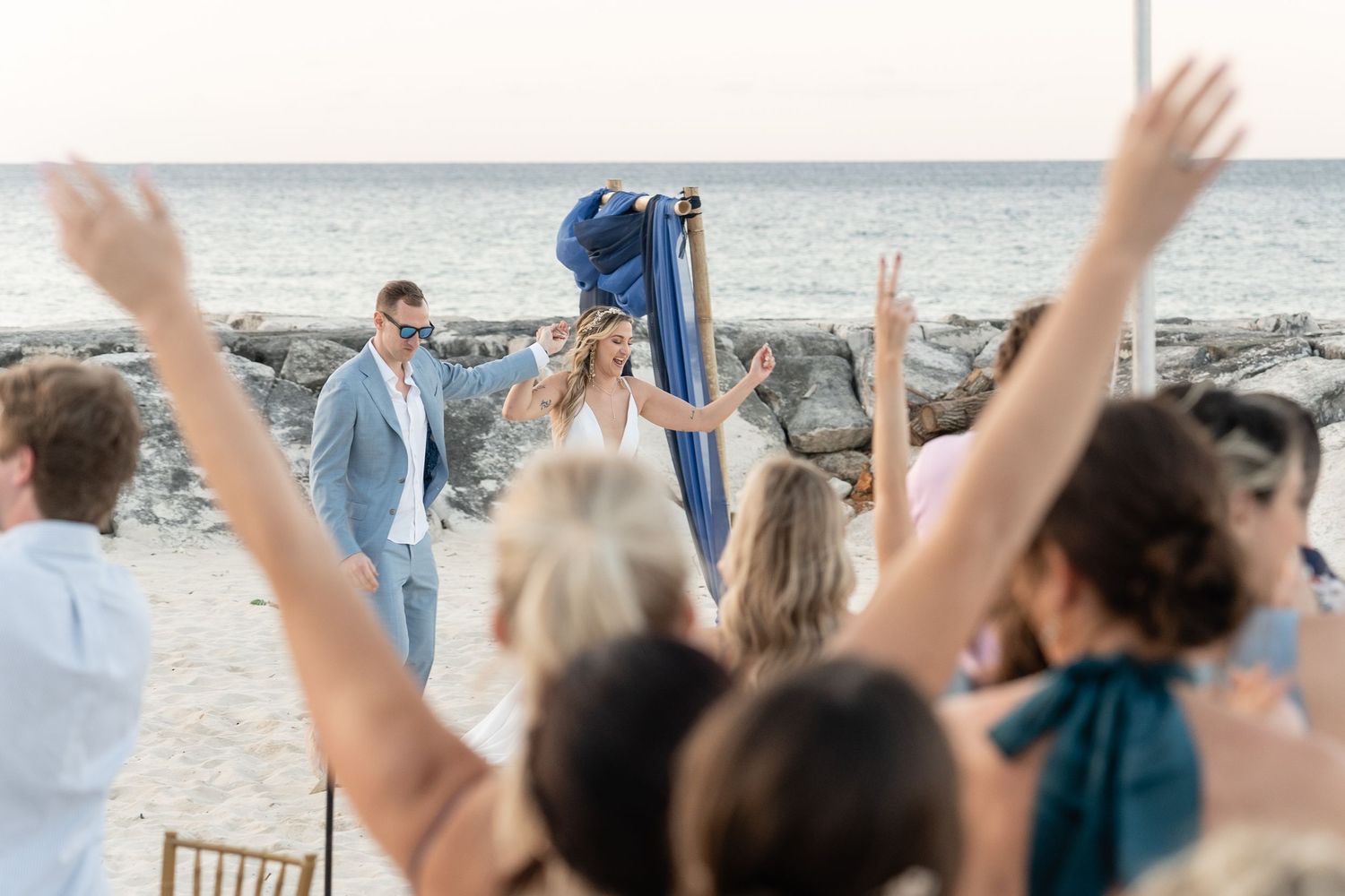 Wedding guests raise arms in celebration during beach ceremony with ocean views.