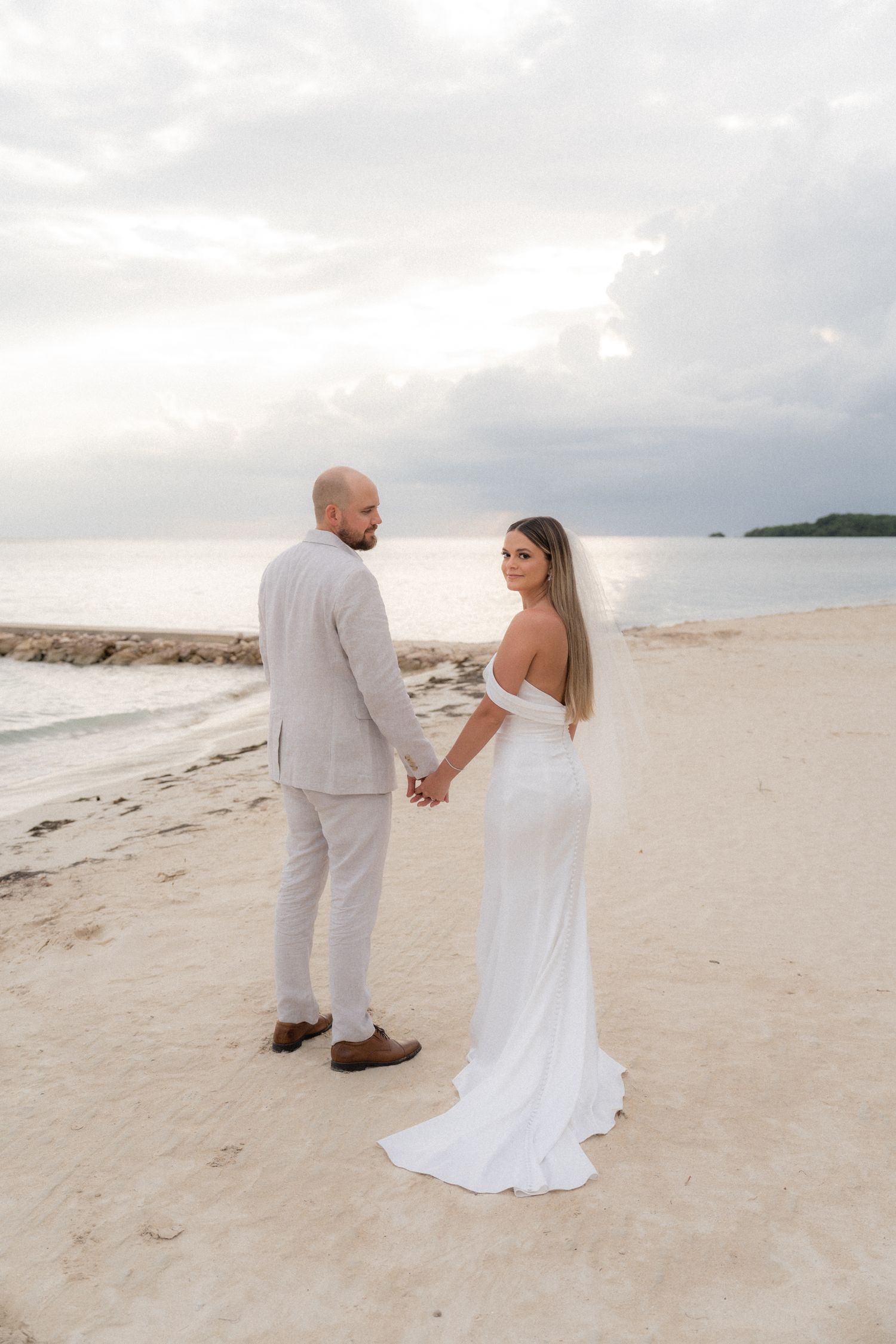 Couple in wedding attire standing on a beach at sunset with ocean waves in the background.