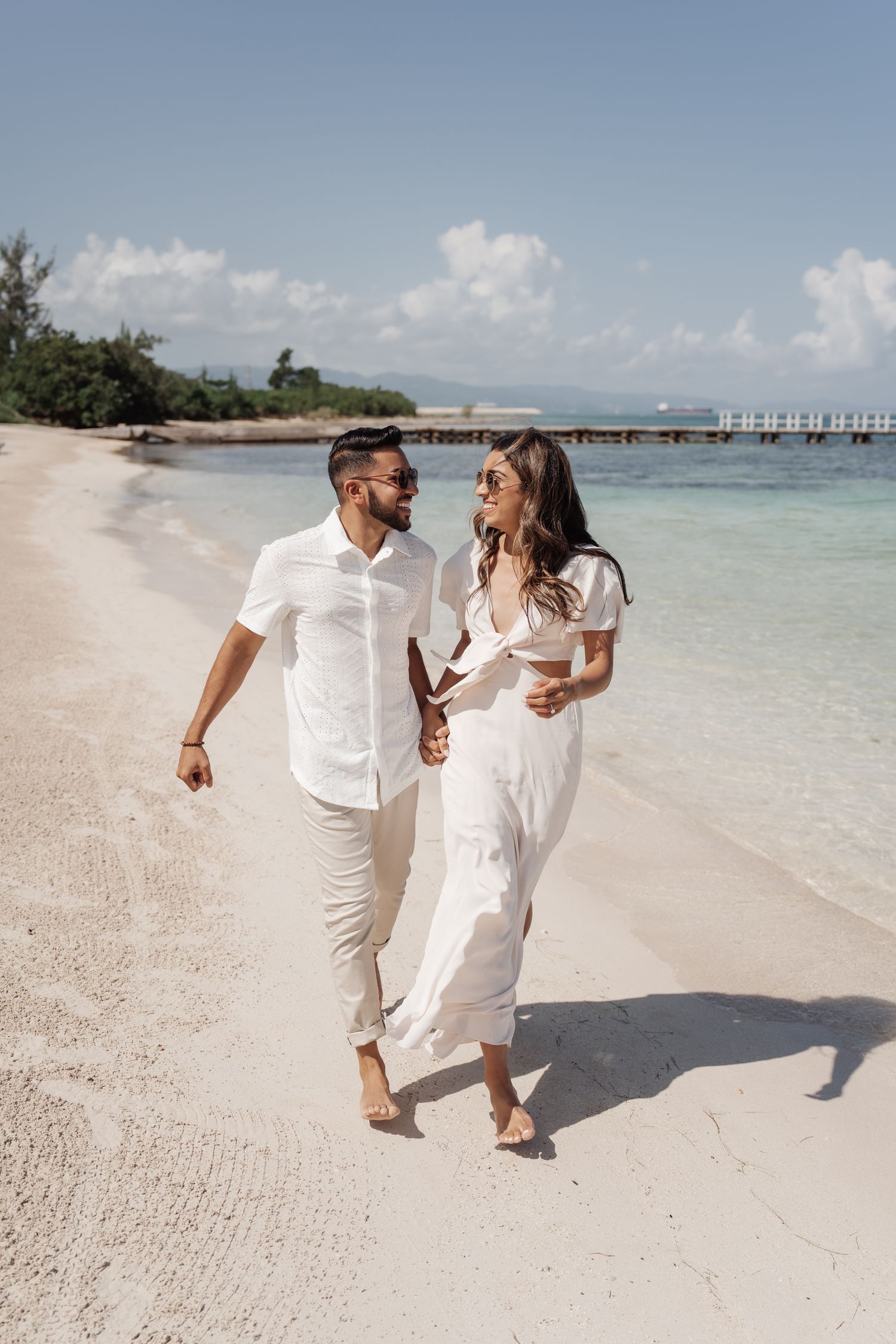 A couple in white outfits walking hand in hand along a scenic tropical beach.