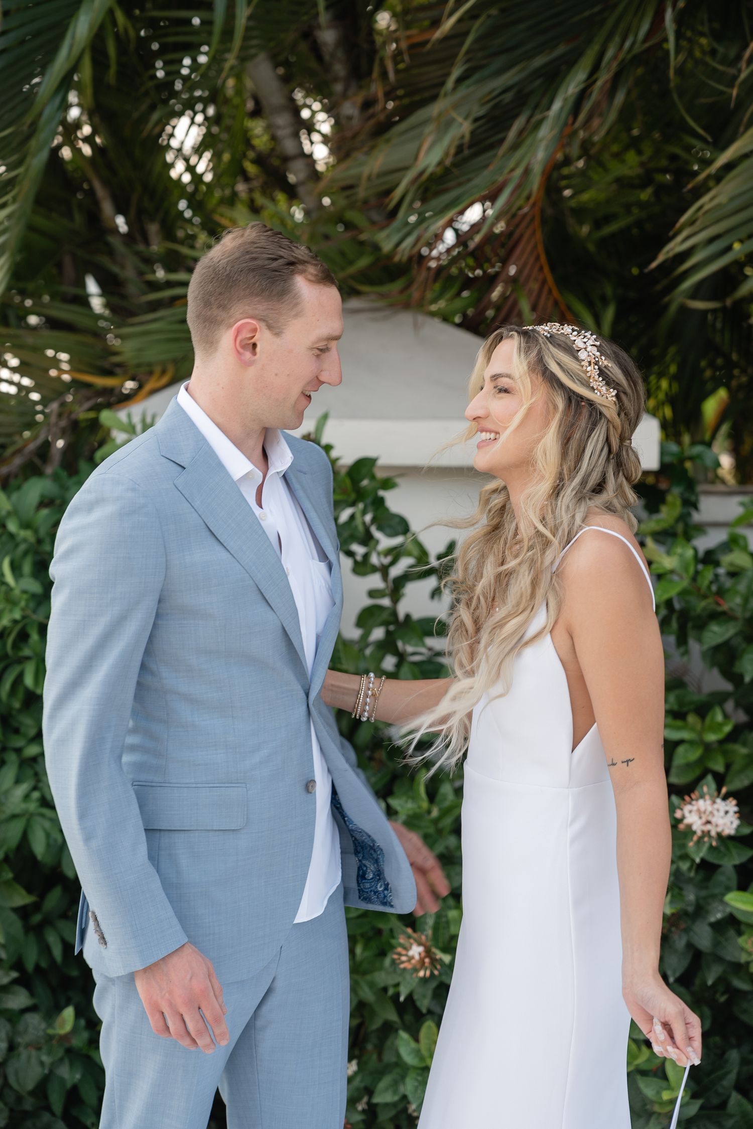 A couple in light blue suit and white dress share a romantic moment among tropical foliage.