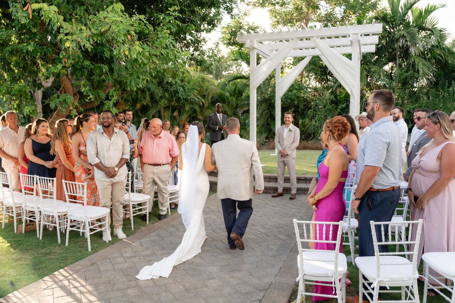 Wedding ceremony recessional with guests seated on white chairs along a garden pathway.