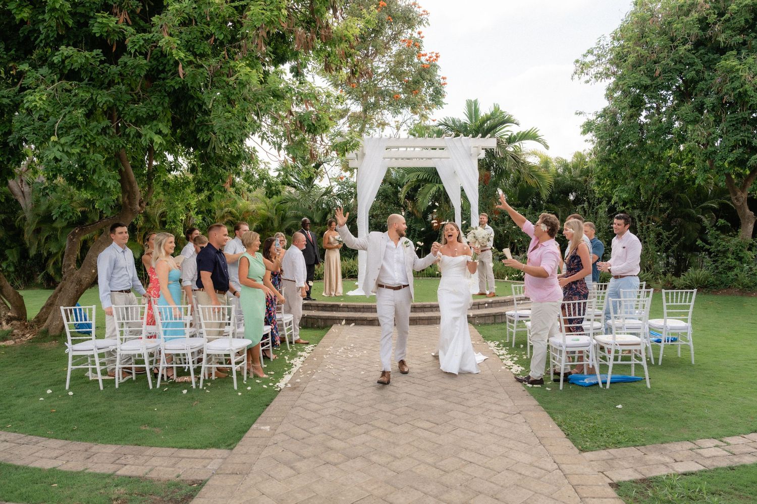 Garden wedding ceremony setup with white chairs and decorated arch on a stone pathway.