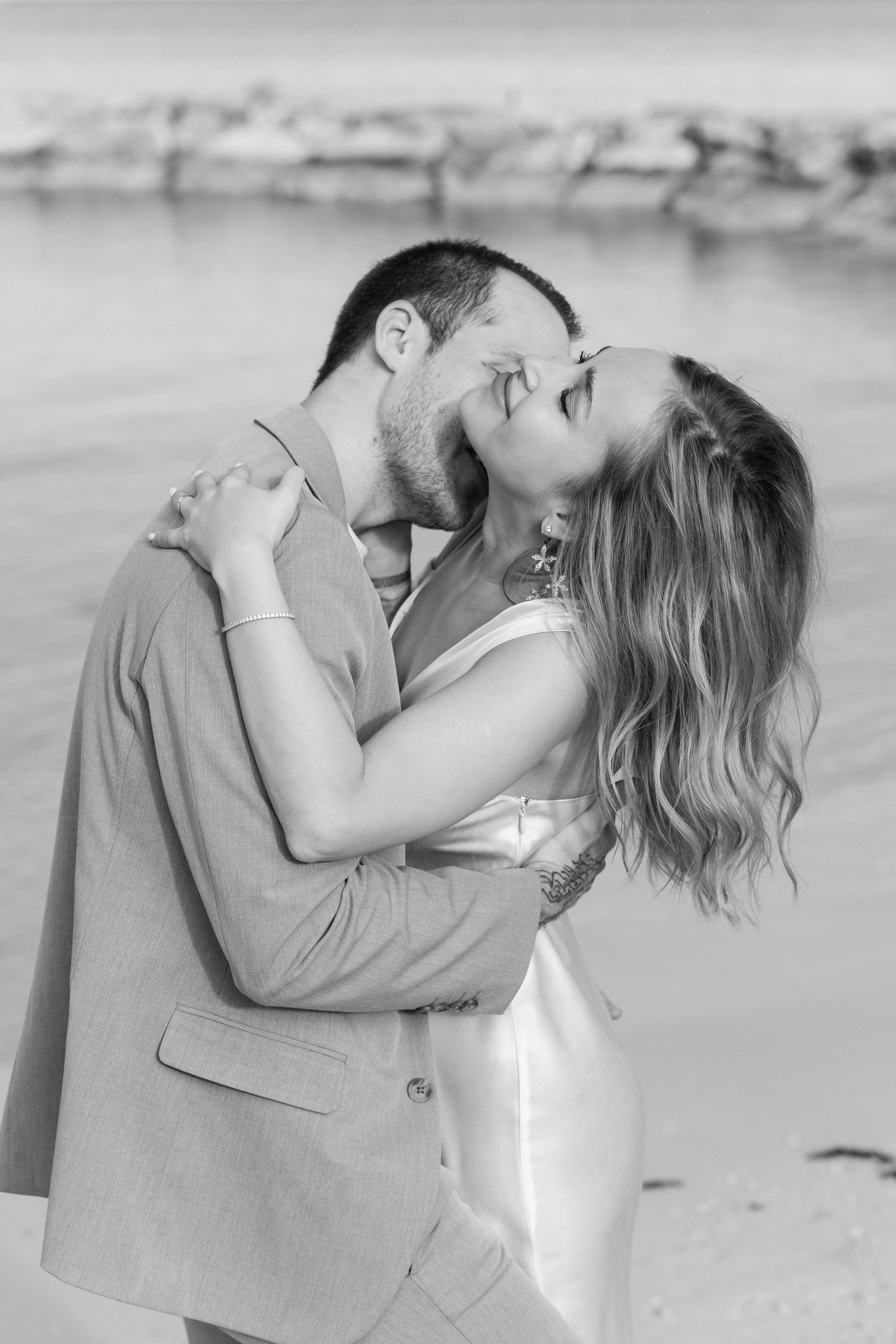 Black and white image of a romantic beach embrace with a stone pier in the background.