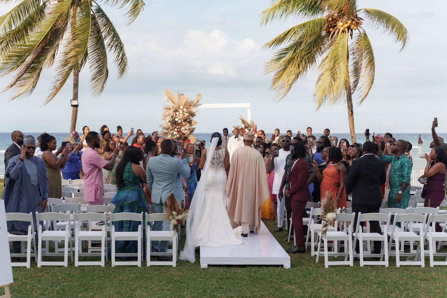 Wedding guests standing for outdoor ceremony under palm trees at tropical beachside venue.