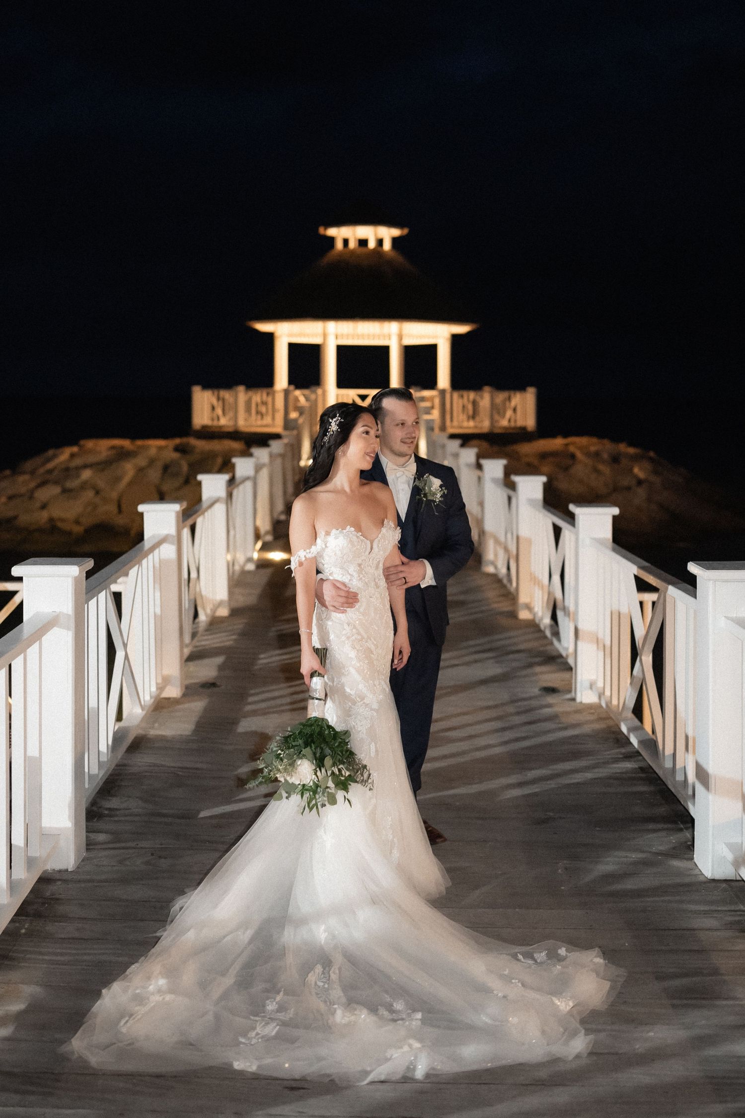 A couple poses on a white wooden pier with a lit gazebo in the background at night.