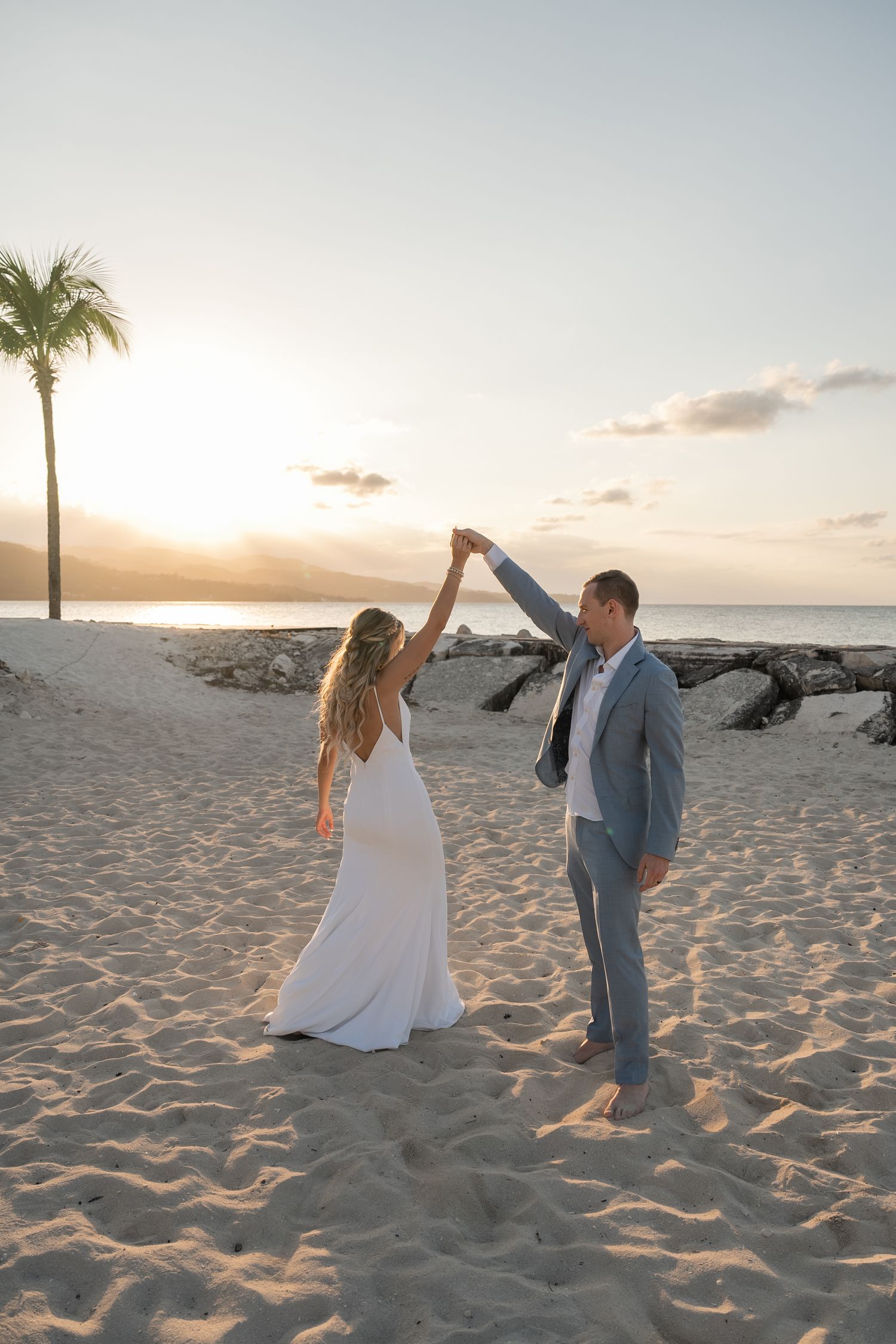 A couple dances on a sandy beach at sunset with a palm tree and ocean view in the background.