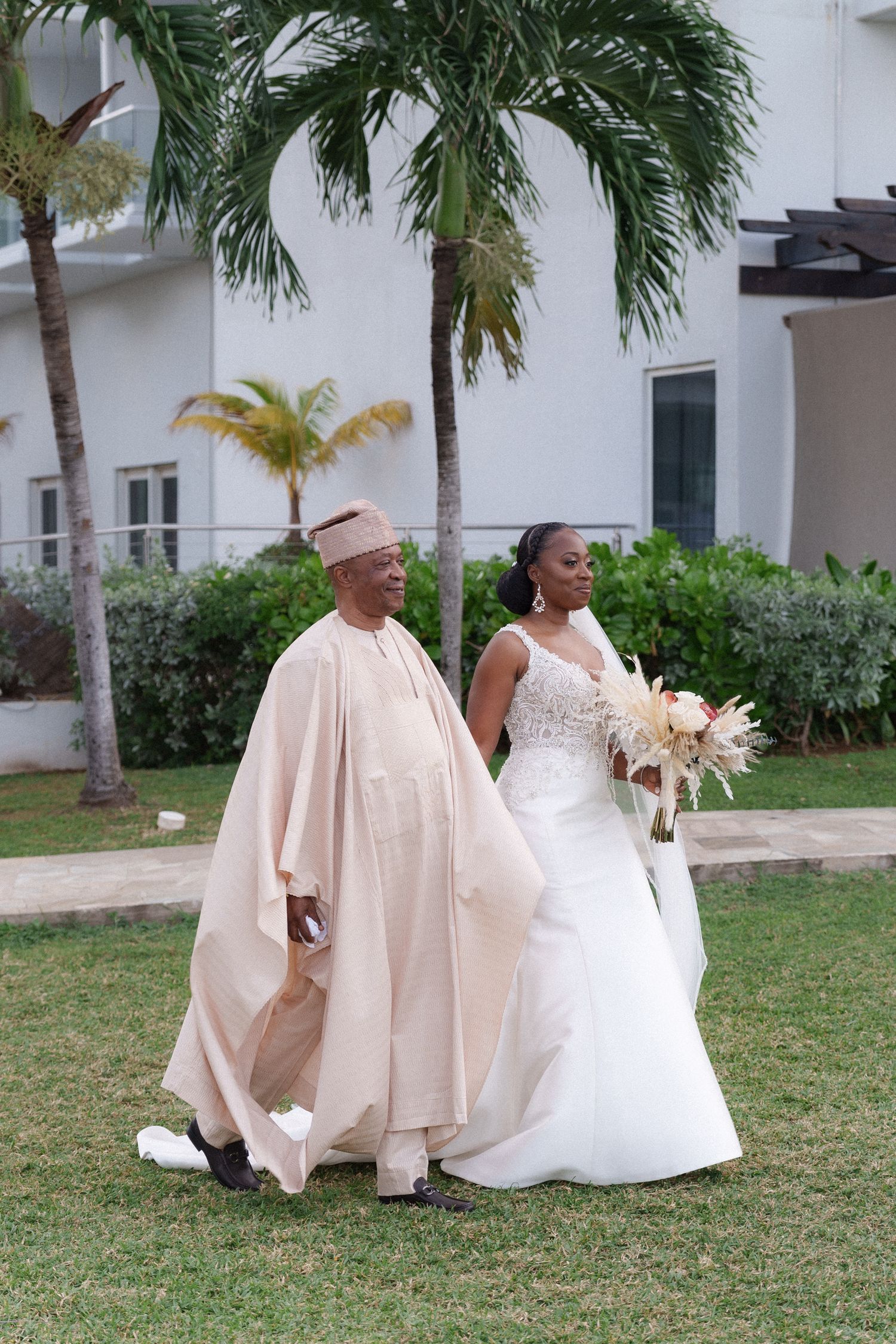 A bride in white gown walking with companion through tropical garden setting.