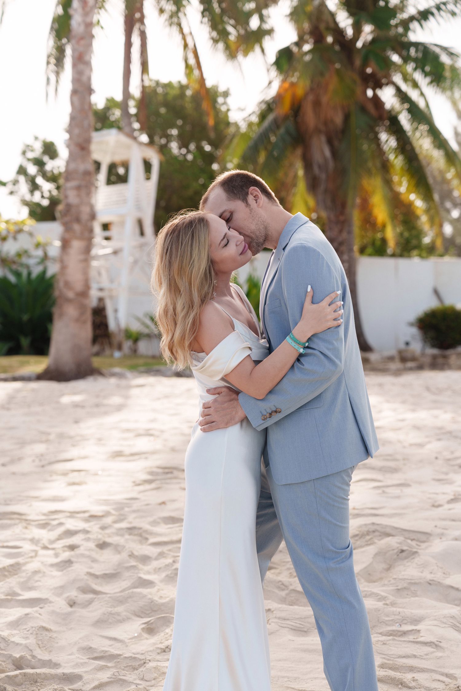 Couple sharing romantic moment near palm trees and white building in tropical setting.